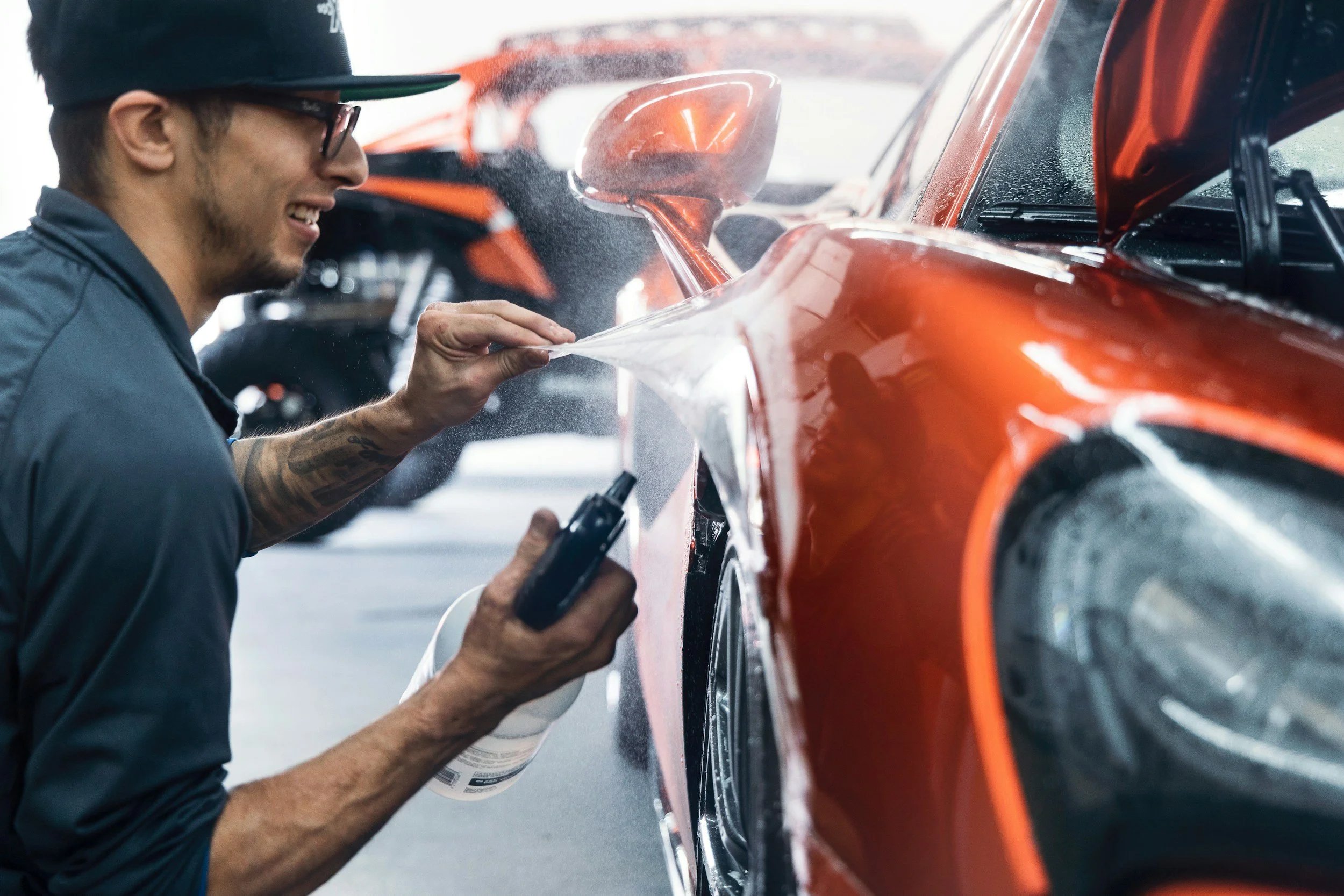 A person wearing glasses and a cap is applying a protective coating or wax to a bright orange car using a cloth, with water droplets on the car's surface.
