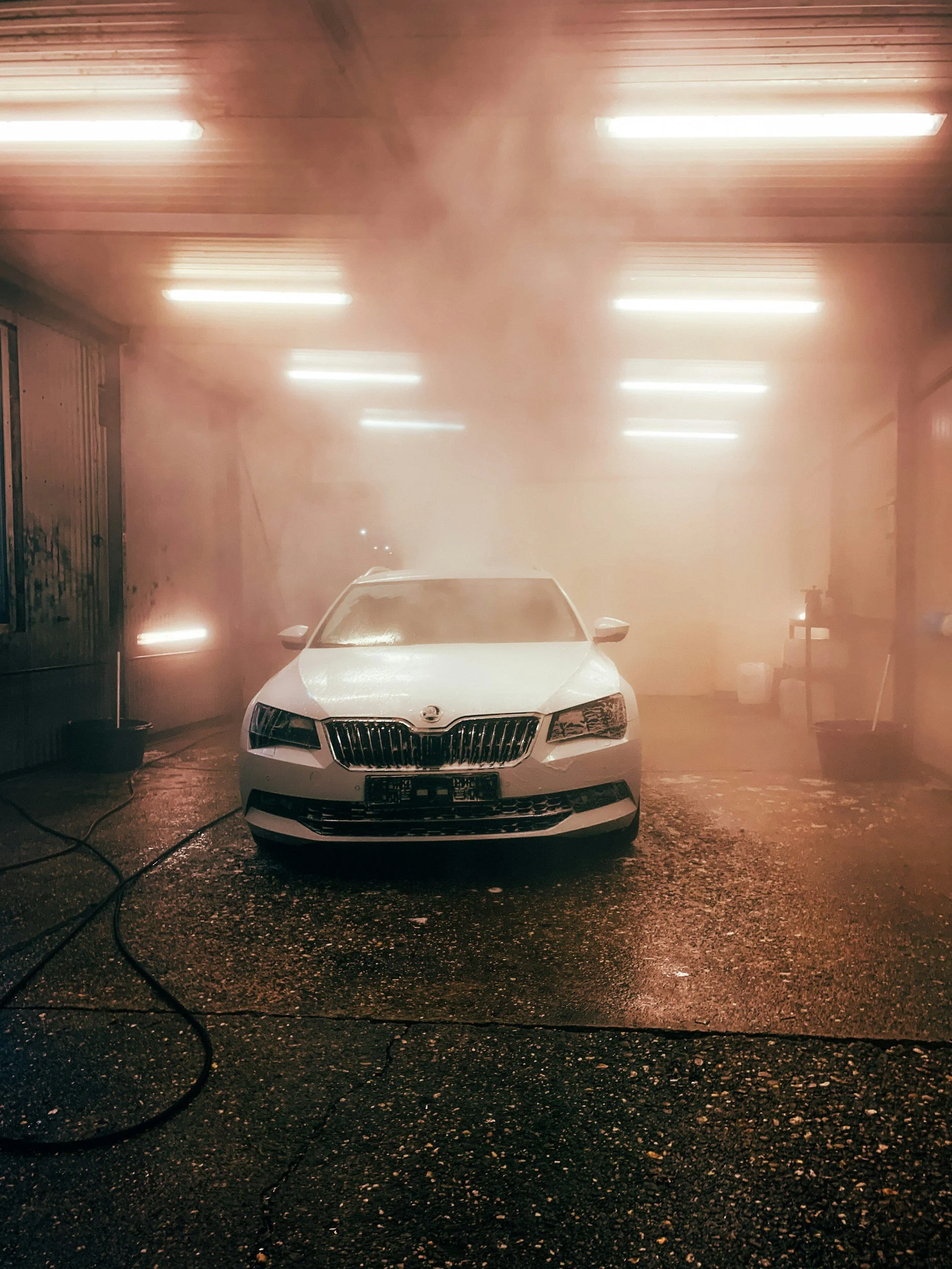 White car parked inside a car wash station with water spray and fog, fluorescent lights on the ceiling, and various cleaning tools on the right side.