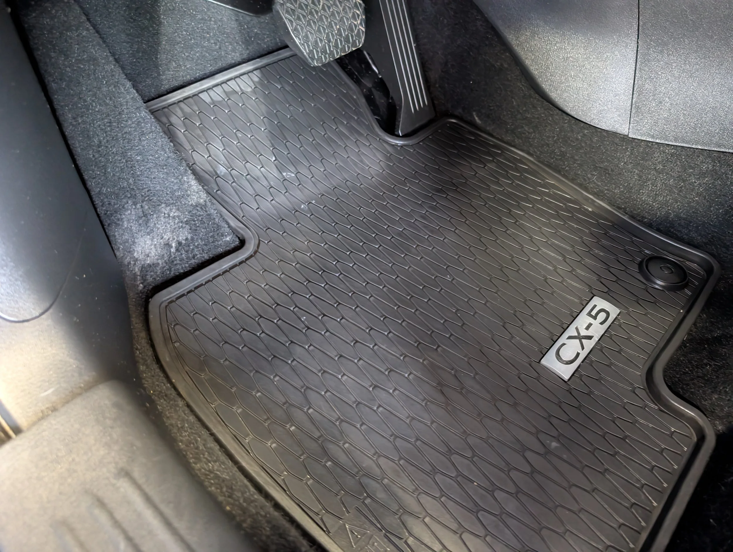 Close-up photo of the car's driver side footwell, showing a black rubber floor mat with a diamond pattern, the gas pedal, and a part of the brake pedal, with some textured carpet around.