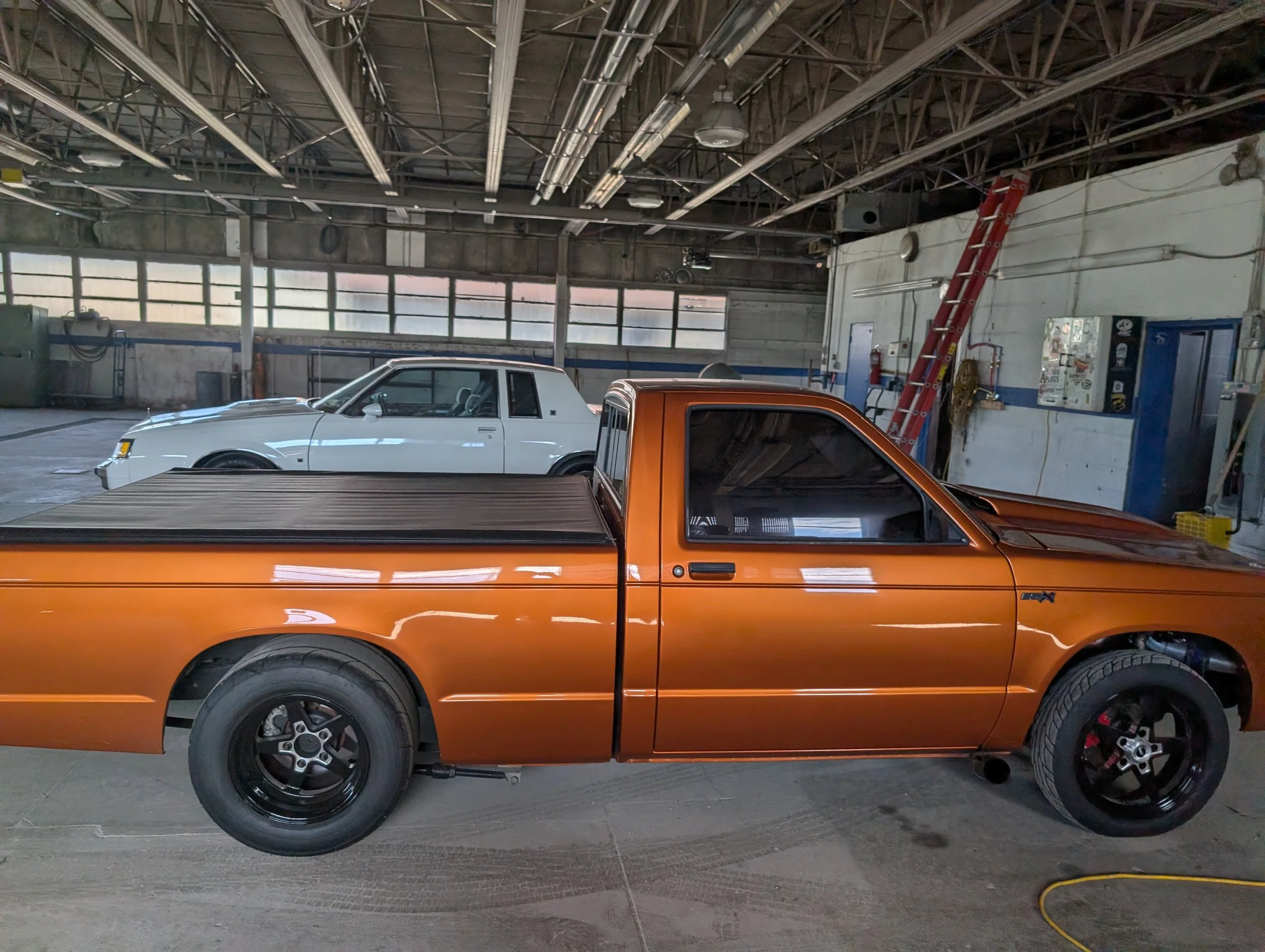 This image shows an indoor garage or workshop with two vintage vehicles, one white and one orange, parked side by side. The garage has a high ceiling with exposed metal beams and several large windows allowing natural light inside. A red ladder is le