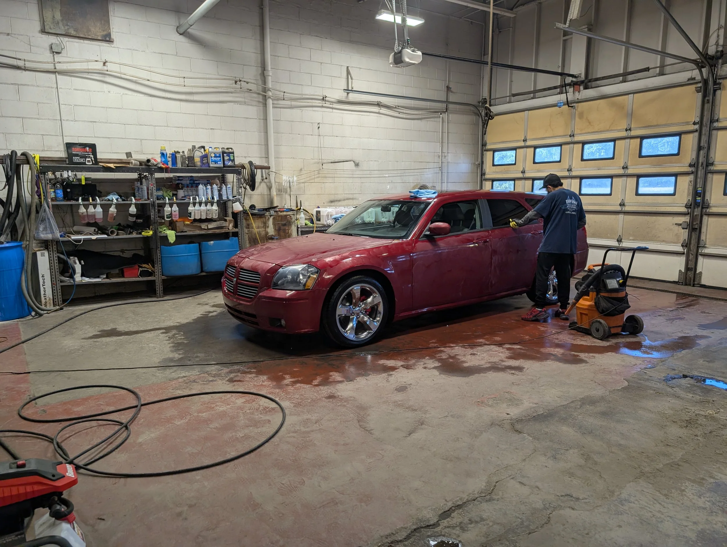 A person working on a red car inside a garage with a yellow and white overhead door, shelves with various tools and supplies, and a wet concrete floor.