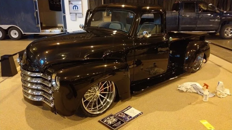 Black vintage pickup truck with custom chrome grille and large multi-spoke wheels on display indoors, surrounded by cleaning supplies and informational signs.