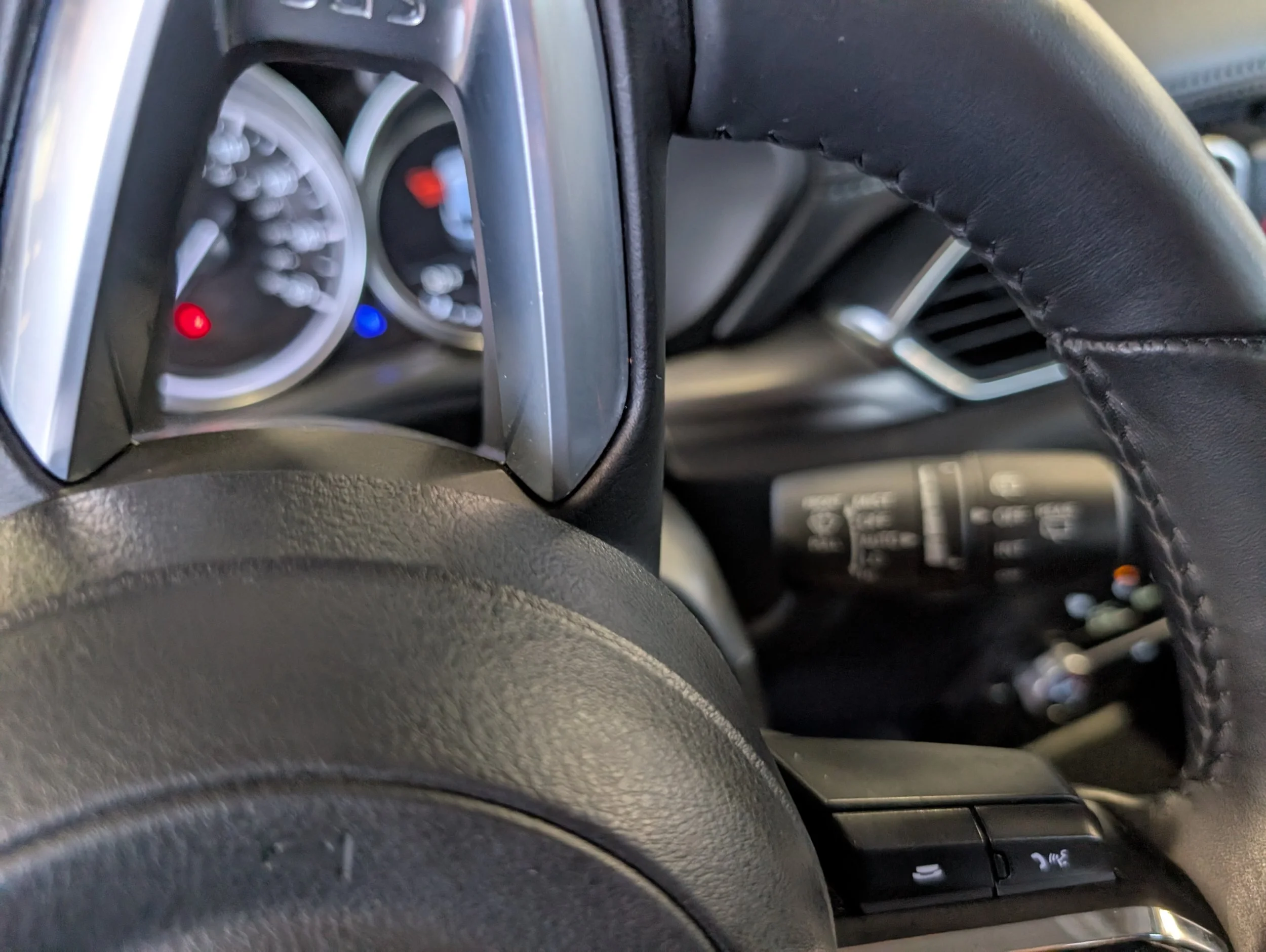 Close-up image of a car dashboard with instrument gauges and control buttons, showing part of the steering wheel covered in black leather.