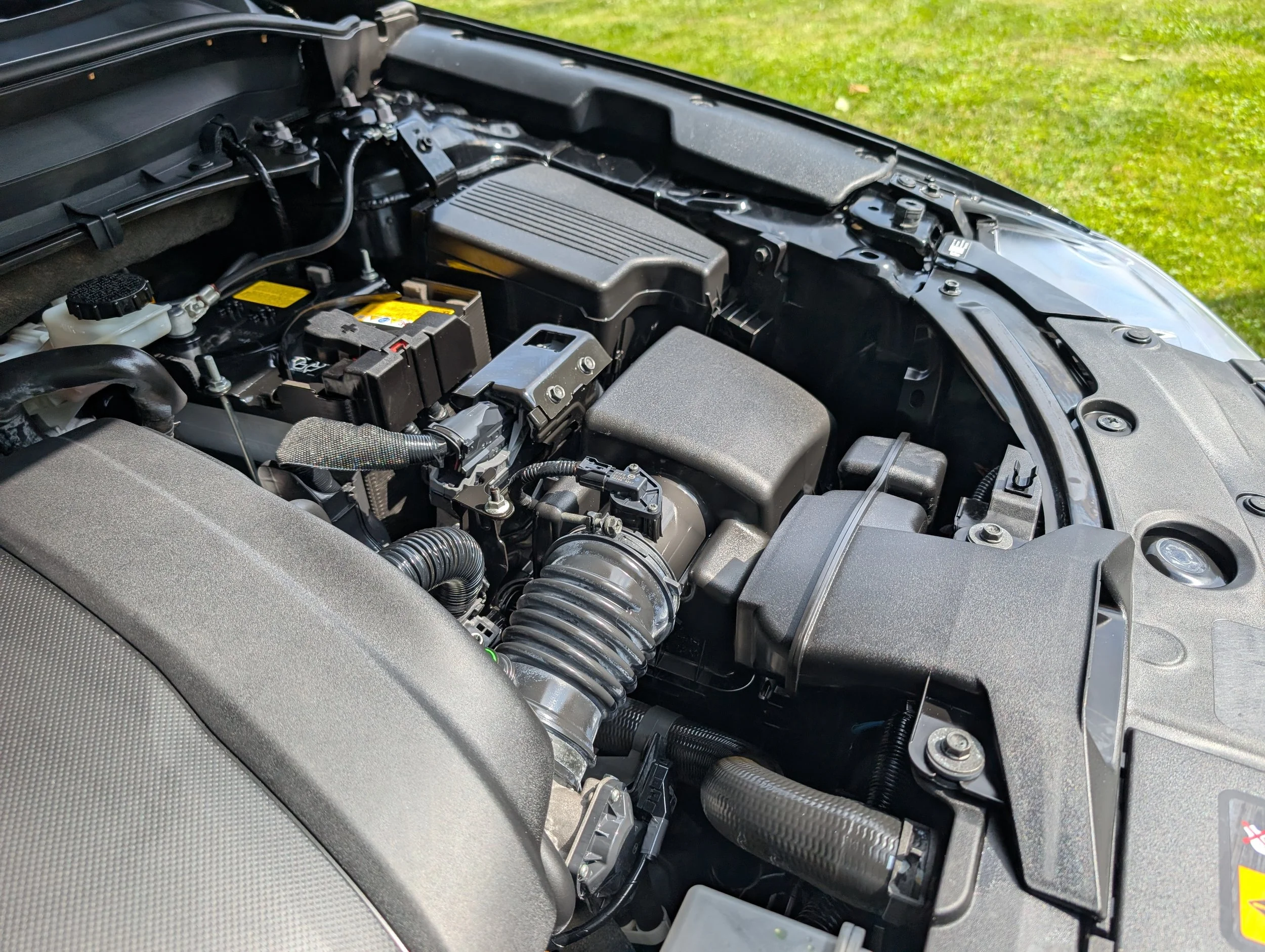Close-up view of a car engine bay showing various components including hoses, battery, and engine cover, with a grassy area in the background.