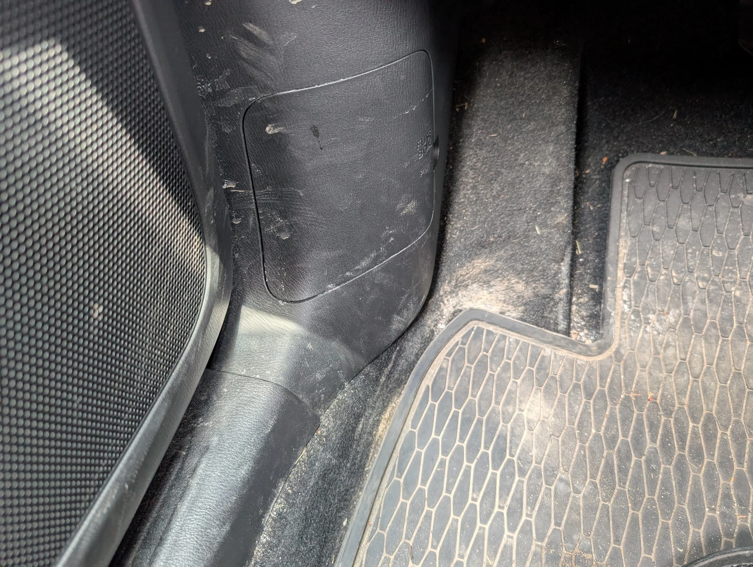 The interior of a vehicle showing a dusty floor mat, a black plastic panel with piece of white string, and a black speaker grille on the left side.