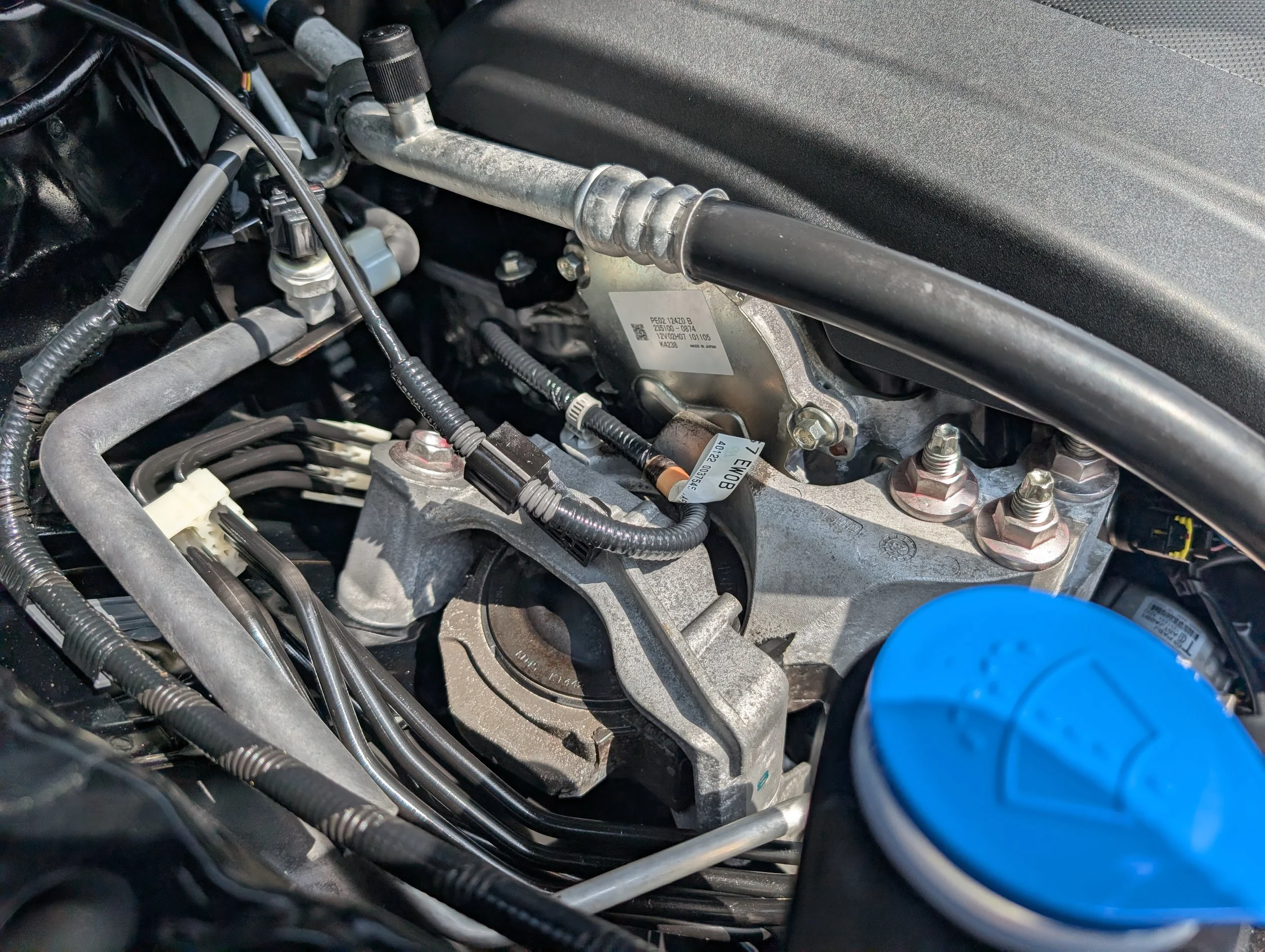 Inner view of a car engine showing various mechanical parts, hoses, and wiring, including a blue wire cap on the right side.