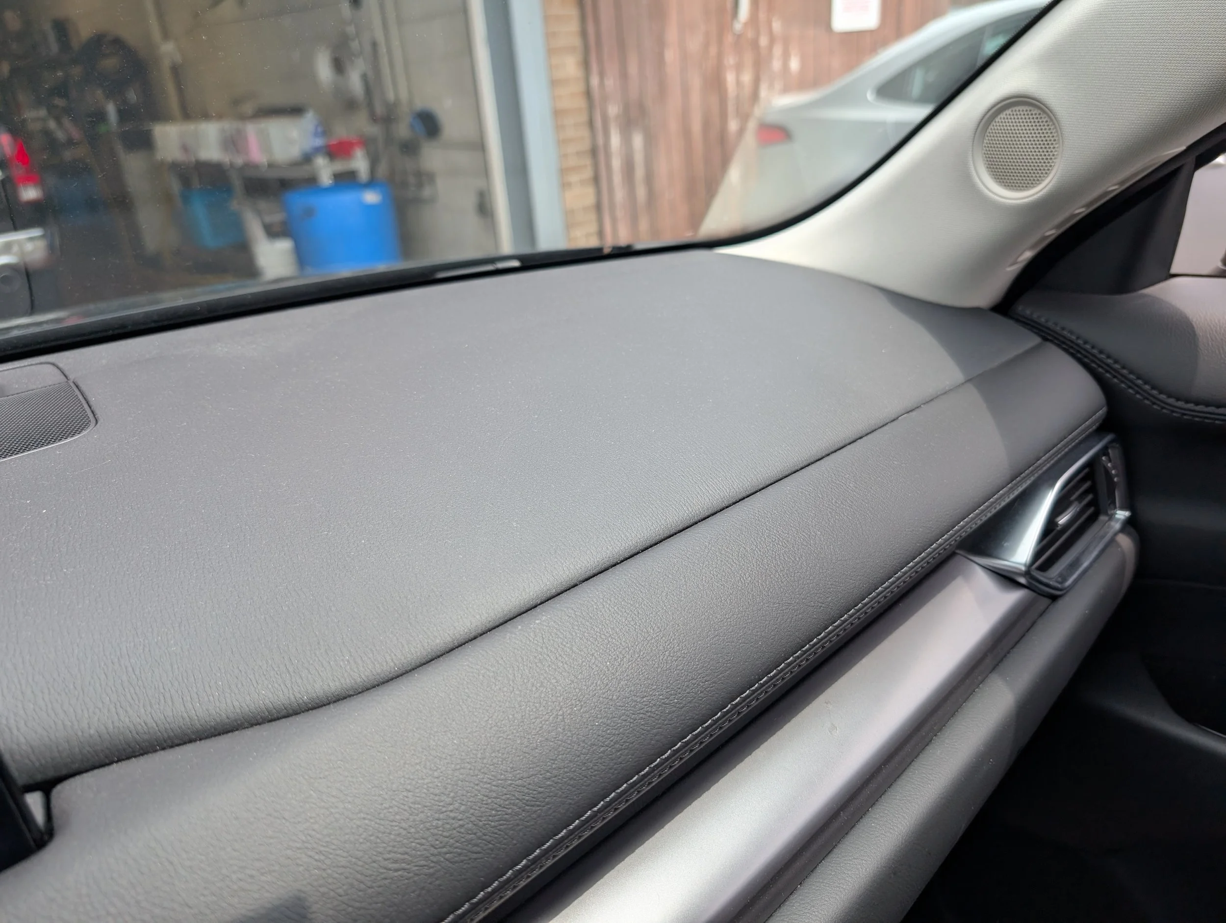 The photo shows the interior of a car with a view of the dashboard, air vents, and part of the windshield. The outside scene through the windshield includes a garage area with a blue barrel, tools, and a brick wall.