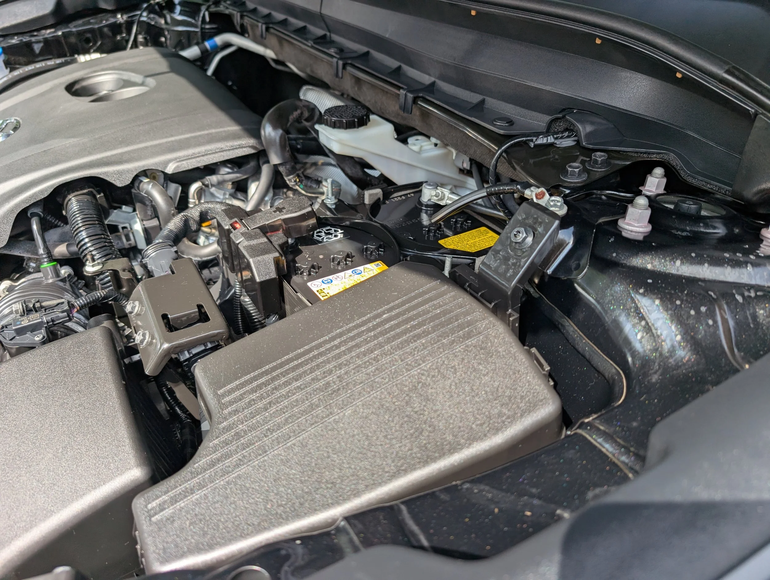 Close-up of a car engine bay showing various engine components, including a plastic coolant reservoir, hoses, and the engine cover.