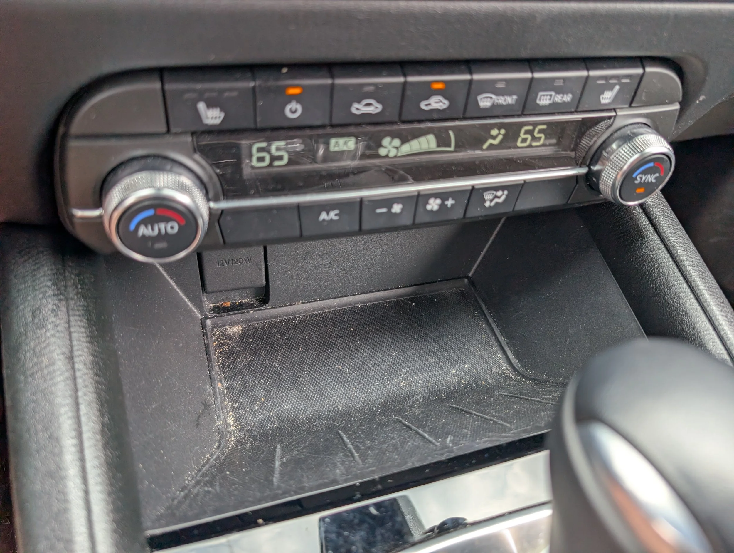 Car climate control panel showing temperature settings at 65 degrees on both sides, with buttons for heated seats, air circulation, defrost, and air conditioning, and a small storage compartment beneath.