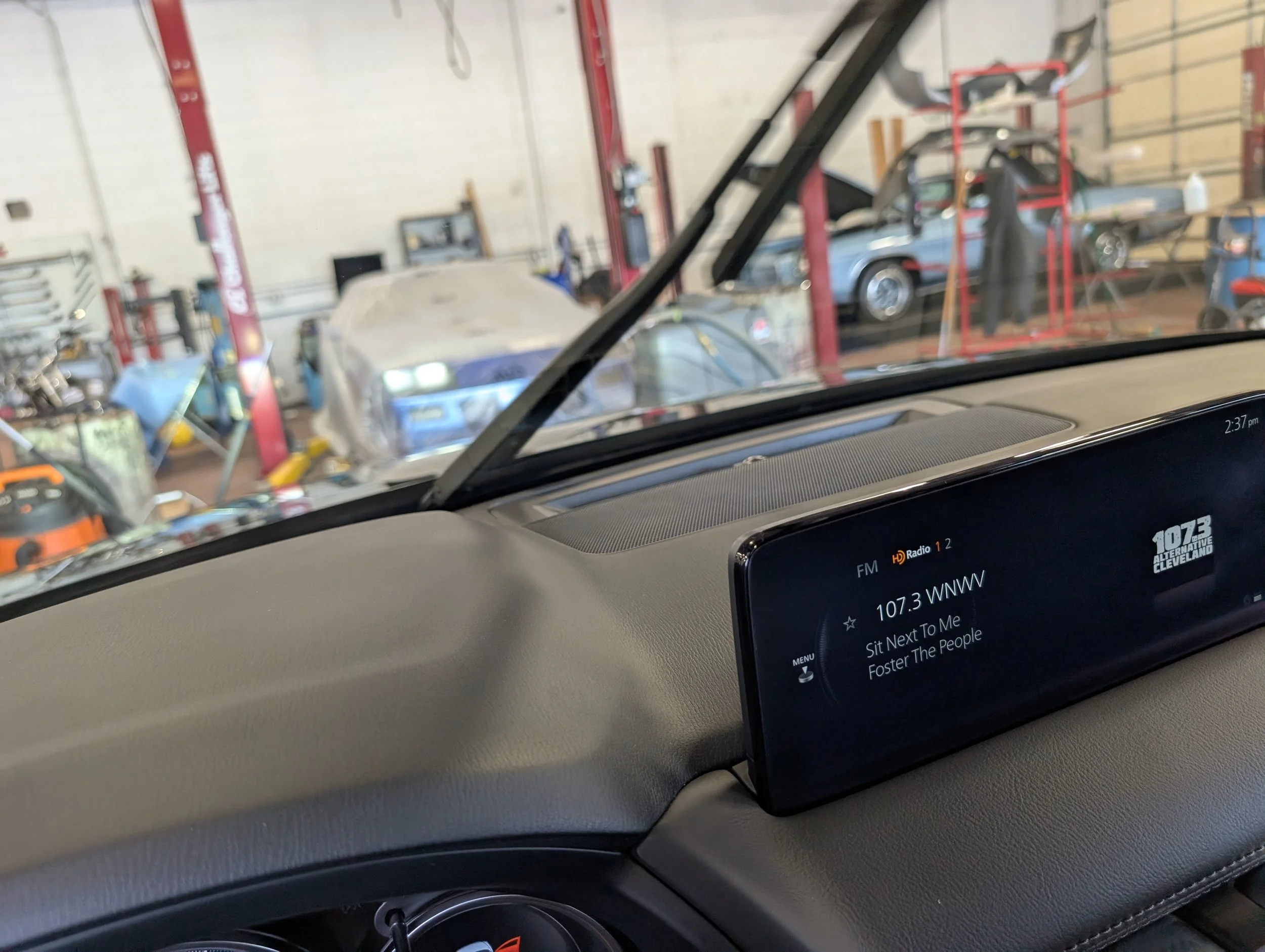 Inside a garage with cars on a lift and tools, viewed through a vehicle's windshield, with a digital radio display showing the station 107.3 FM.