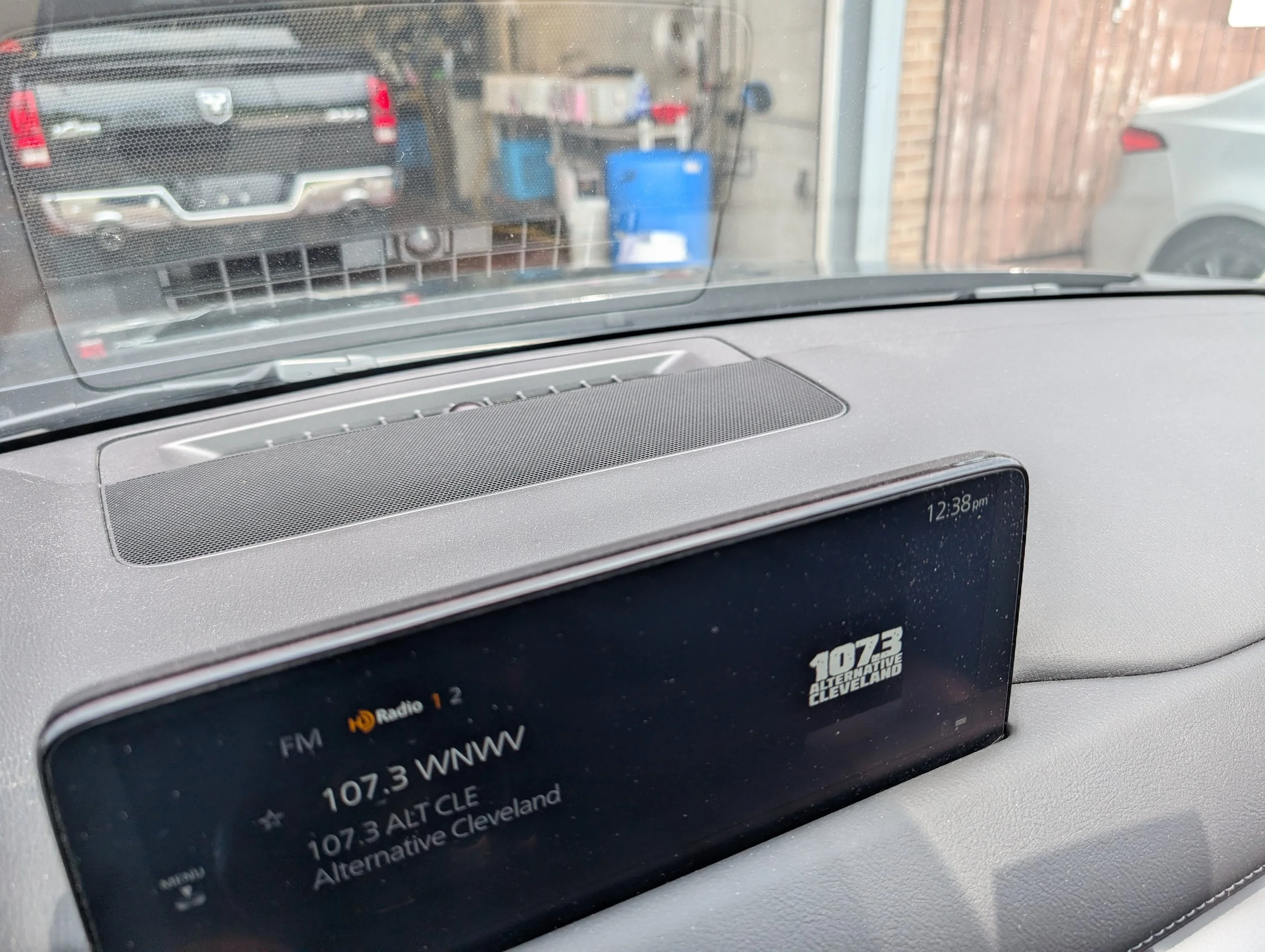 Car dashboard display showing radio station 107.3 alternative Cleveland, with the timestamp 12:38 pm. The view through the windshield shows a garage with a vehicle, a blue container, and a brick wall outside.