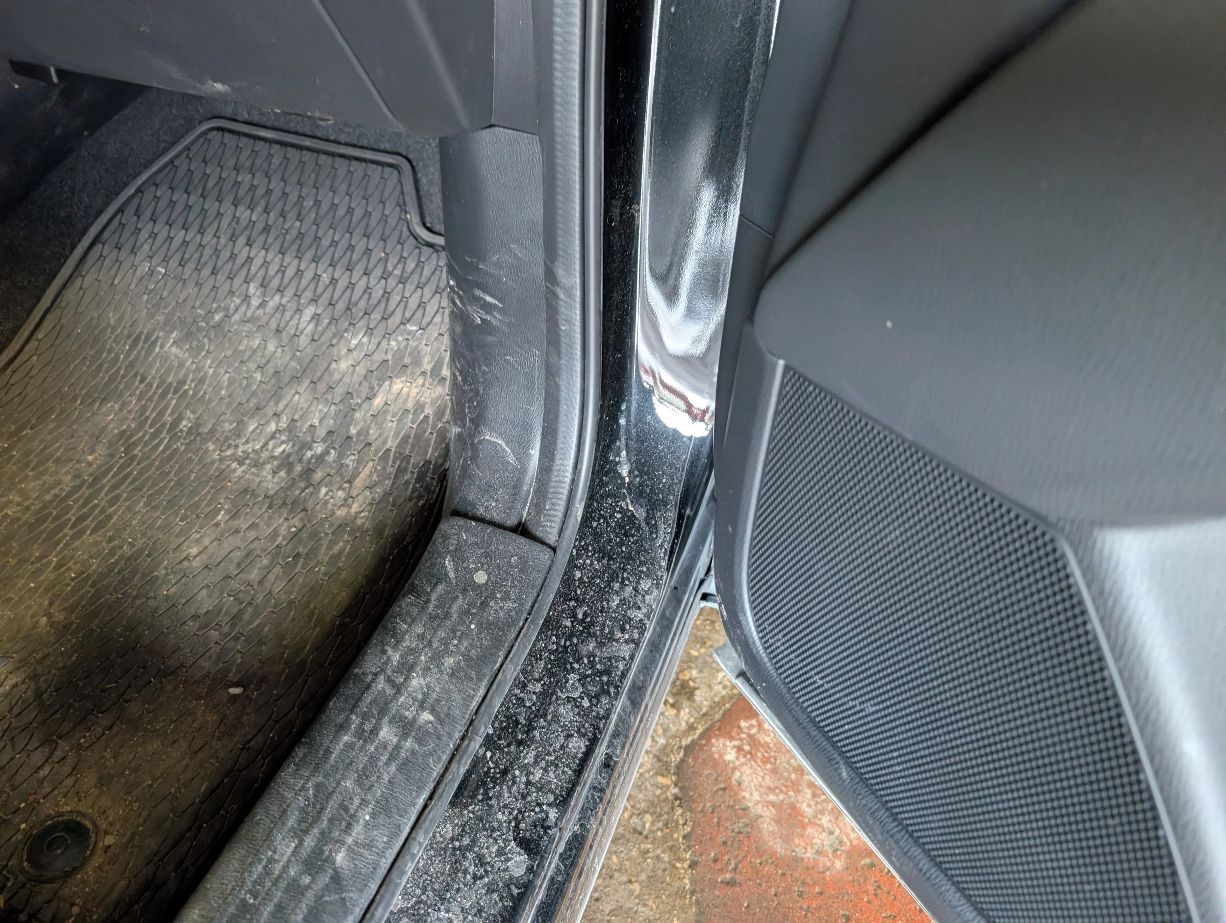 Close-up of the side of a black car with a sliding door open, showing the interior floor mat and door sill with some dirt and scuff marks.
