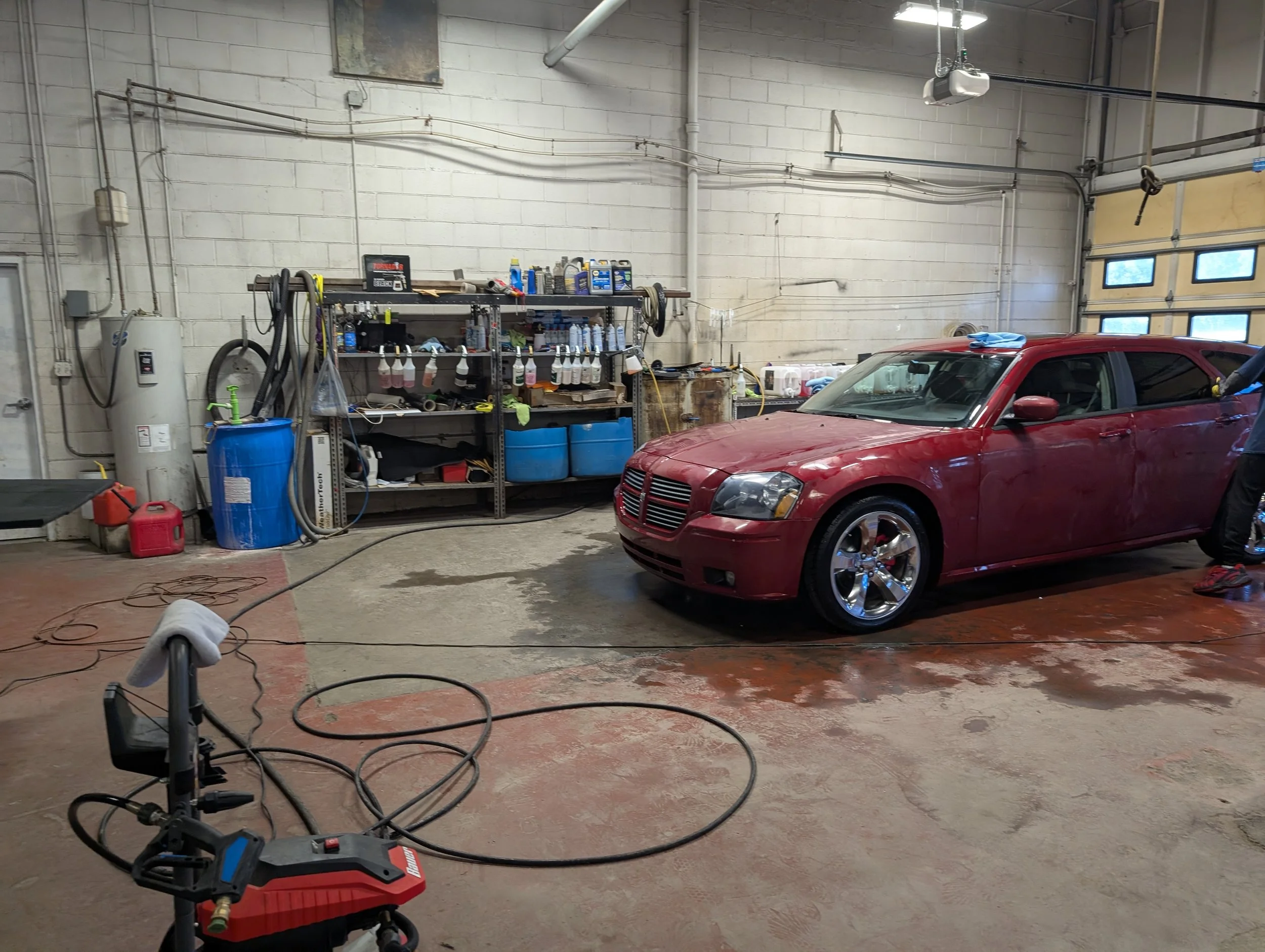 An indoor garage scene with a red car parked near a workbench. The garage walls are white cinder blocks, and various tools, cleaning supplies, and equipment are on shelves and in containers. There are hoses, a blue barrel, and a cleaning machine on t