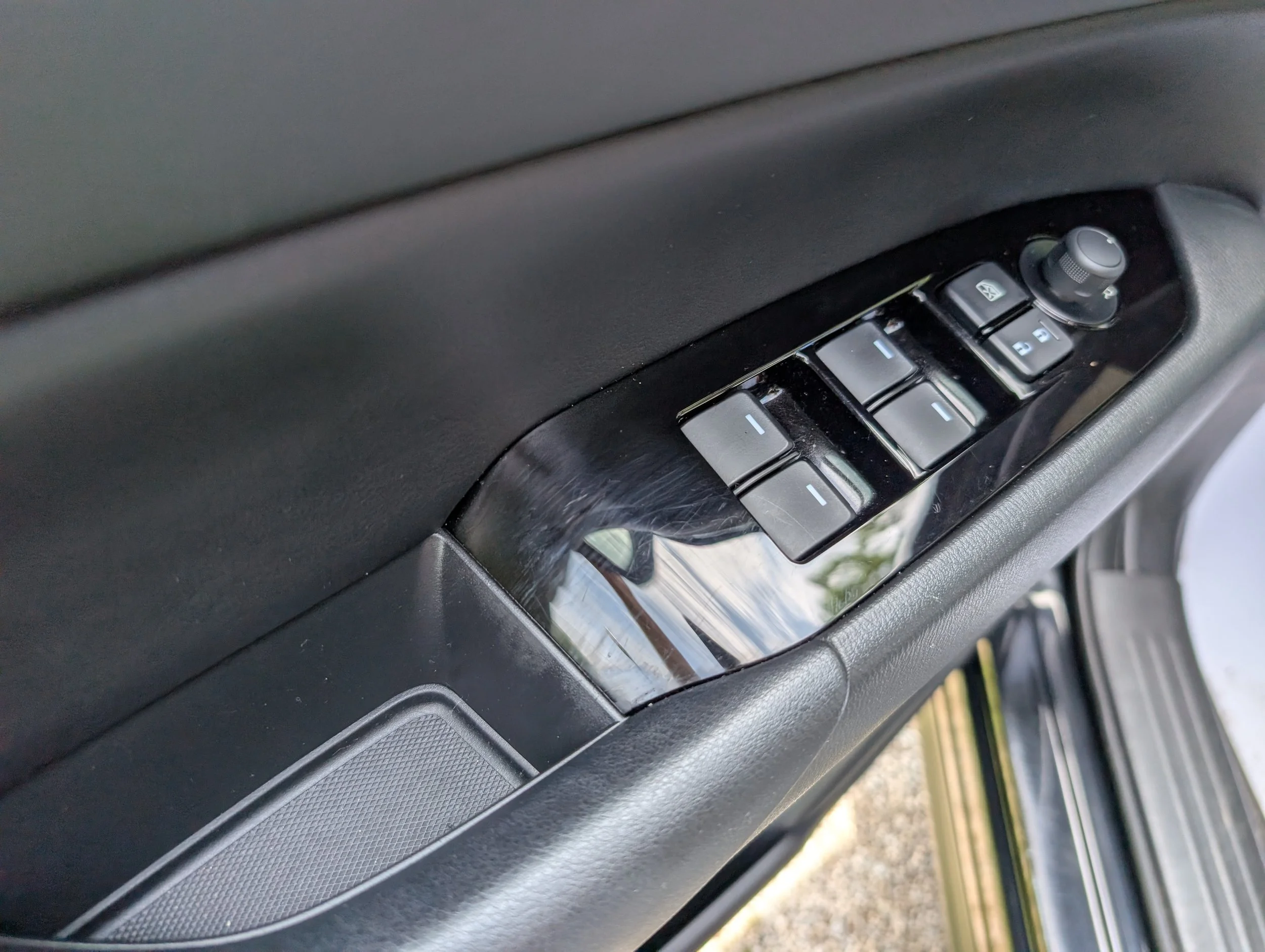 The image shows a close-up view of the driver side door panel of a car, including the window control buttons, lock/unlock buttons, and side mirror adjustment knob.