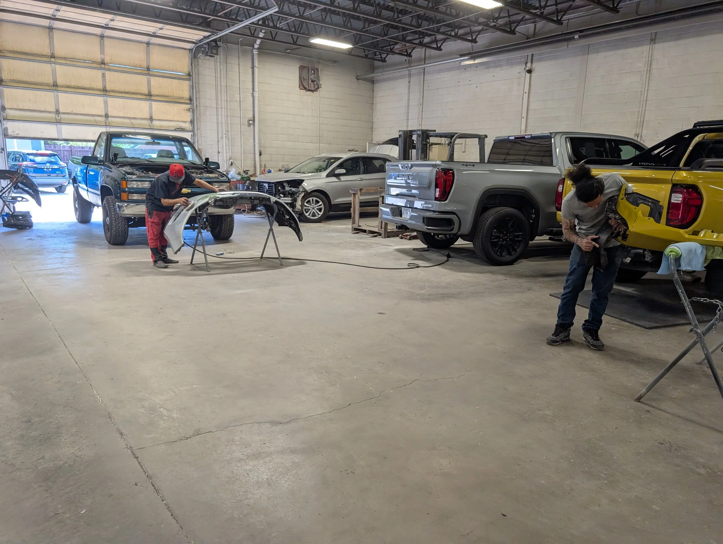 Inside an auto repair shop with several cars being repaired or restored, including a yellow truck and a silver truck, with two mechanics working on different vehicles.