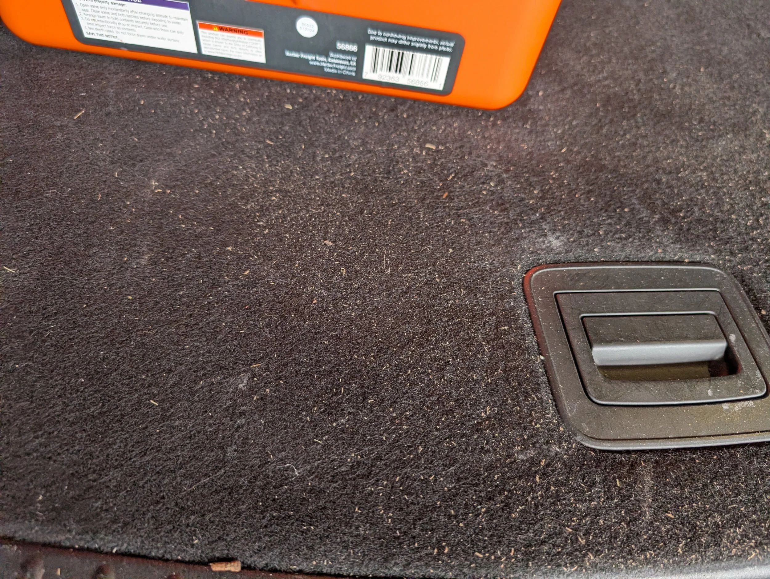 Close-up view of a black vehicle's interior floor mat with an orange object partially visible at the top and a black rectangular vent or button at the bottom right corner.