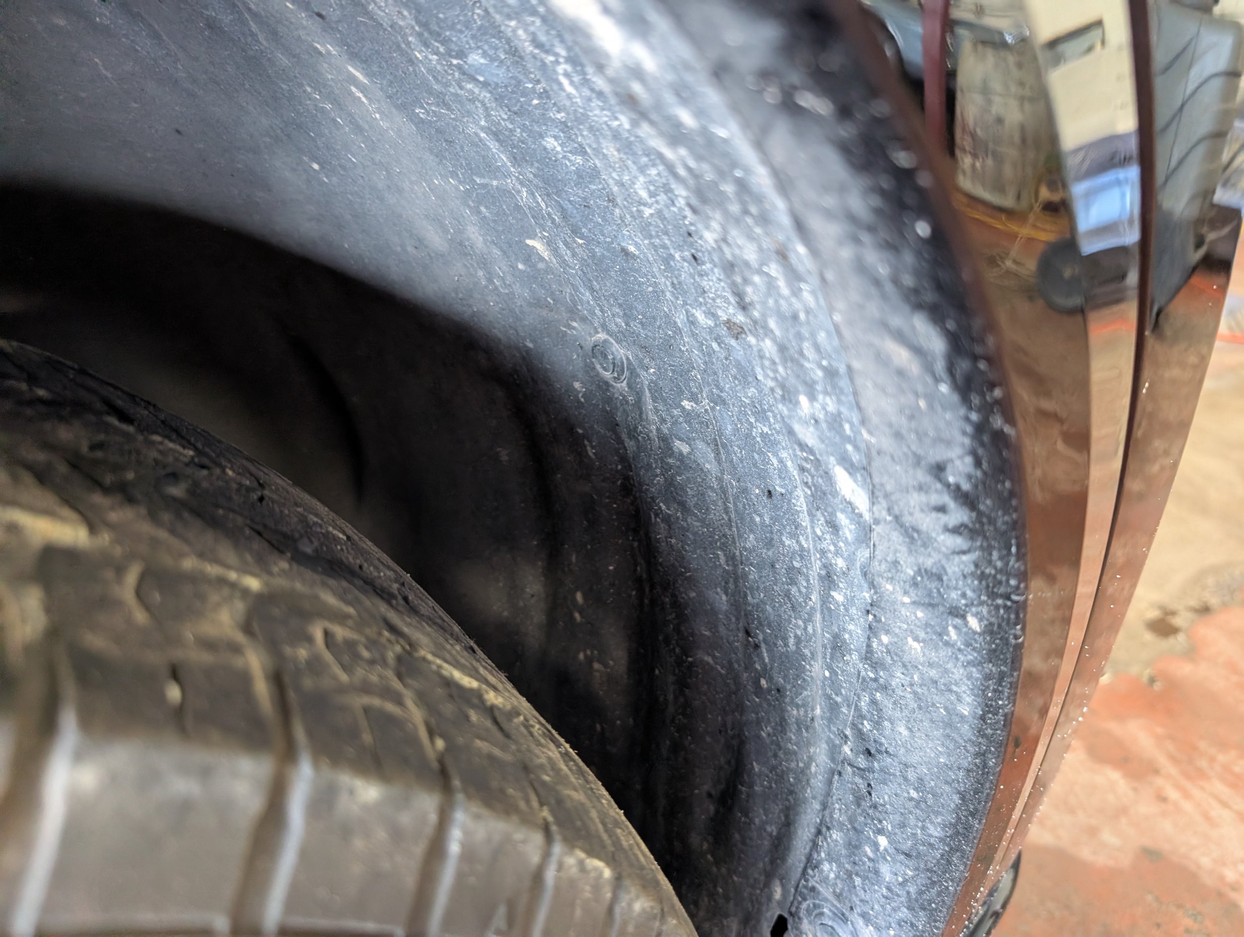 A close-up view of a dirty, dusty vehicle wheel well showing the edge of a tire, with a small portion of the car’s body and paint reflected on a nearby metal part.