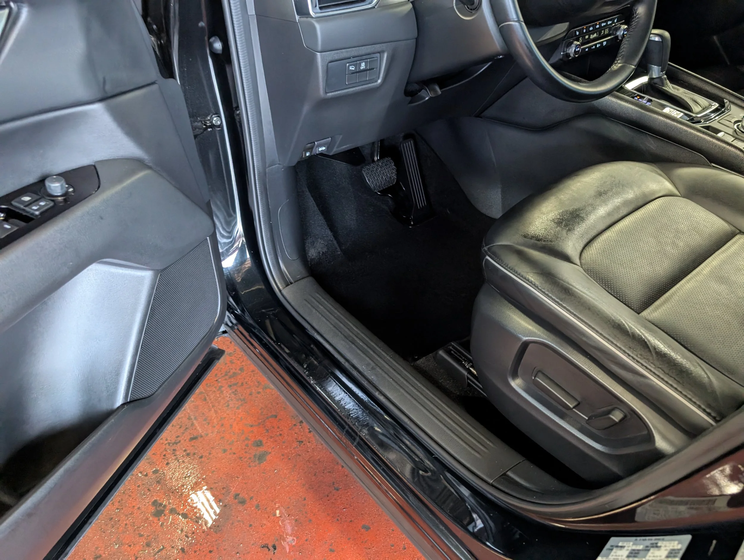 Inside view of a car showing the driver's side, including the black leather seat, steering wheel, dashboard, accelerator and brake pedals, and part of the center console.