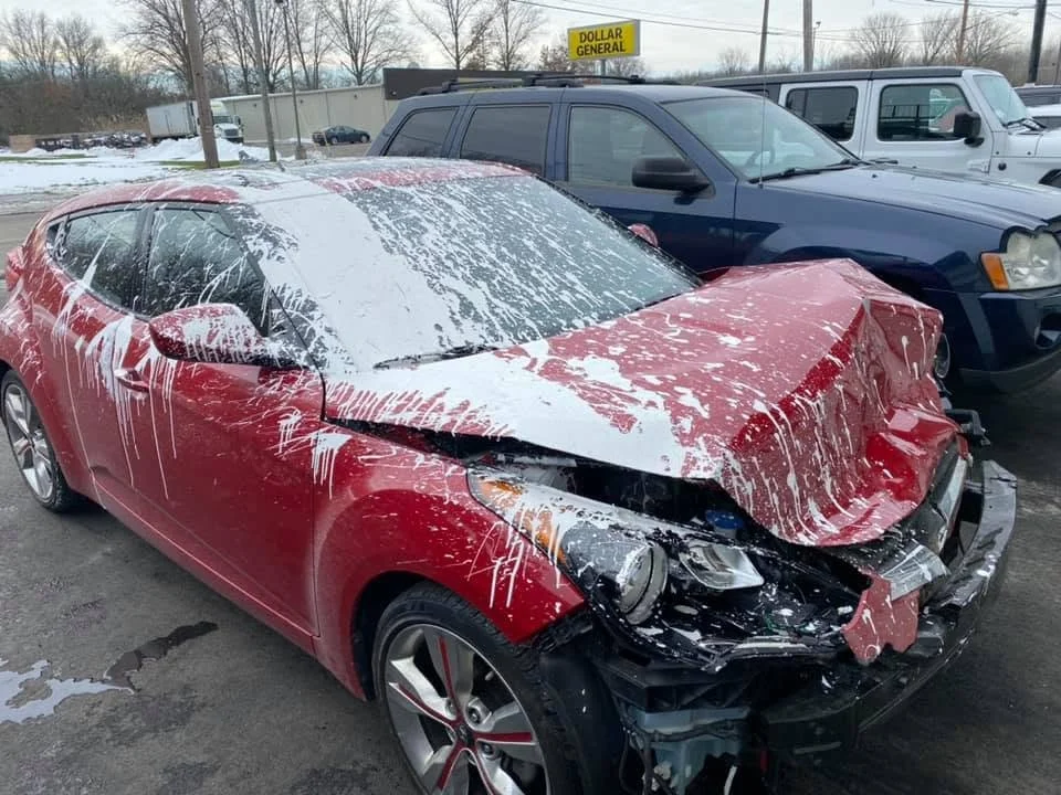 Red car with significant front-end damage and foam cleaning or snow on the windshield, parked in a lot next to black and white vehicles, with a Dollar General store in the background.