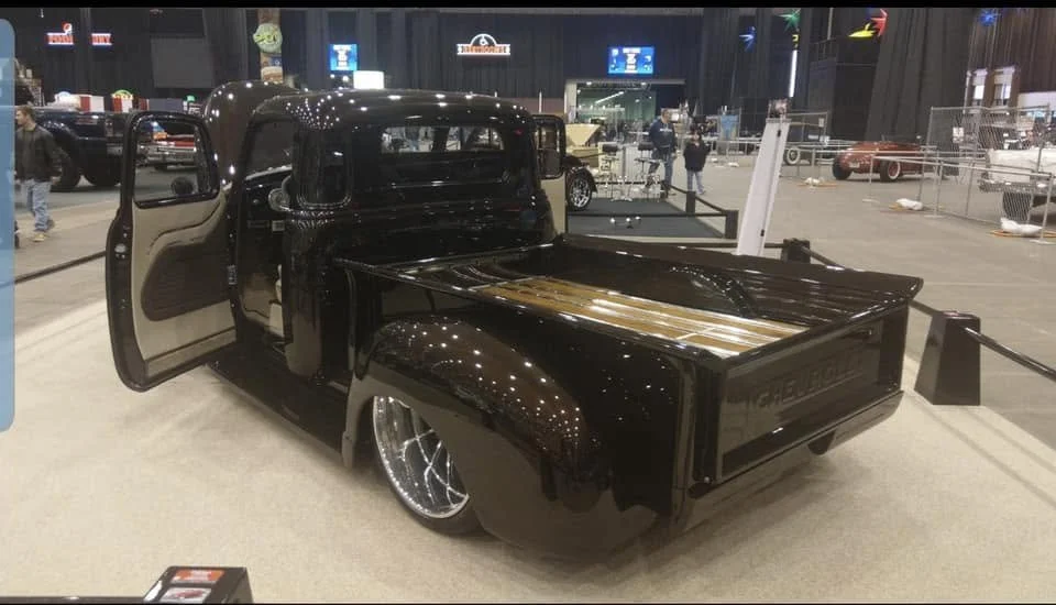 A vintage black pickup truck with a wooden flatbed on display at a car show, with its door open and shiny silver wheels.