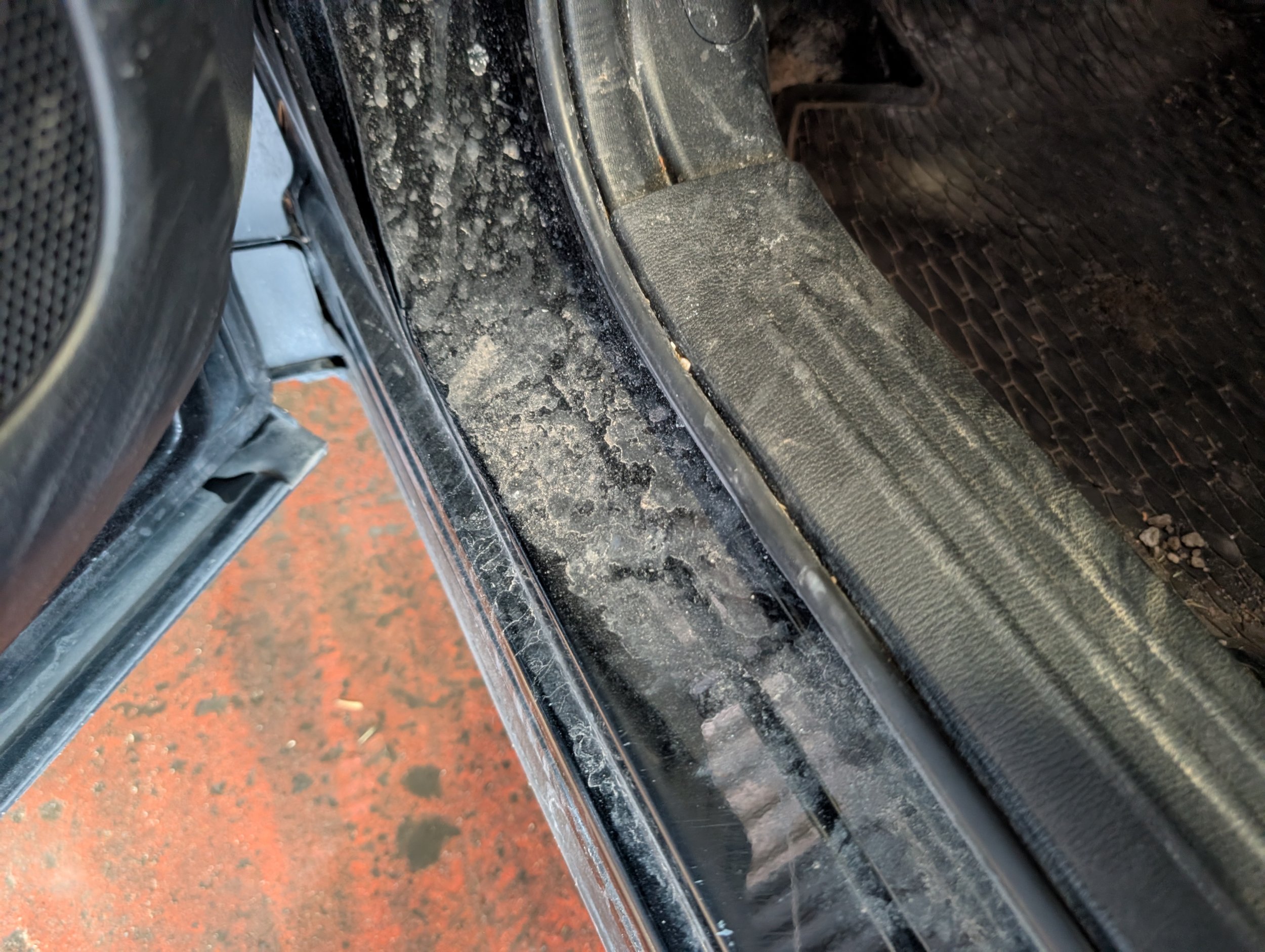 A close-up view of a dirty and dusty car door sill, with a portion of the vehicle's black interior and rubber mat visible.