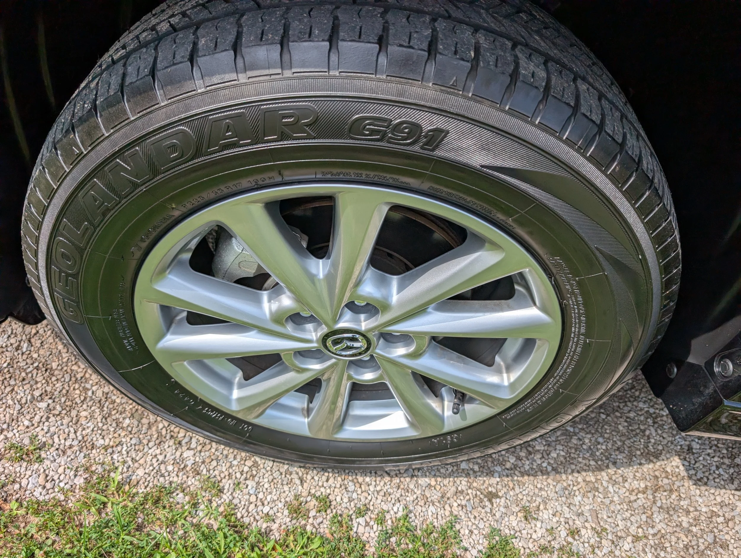Close-up of a car tire with the sidewall displaying the tire model 'Touring A/S G91' mounted on a silver alloy wheel, resting on gravel with some green plants nearby.