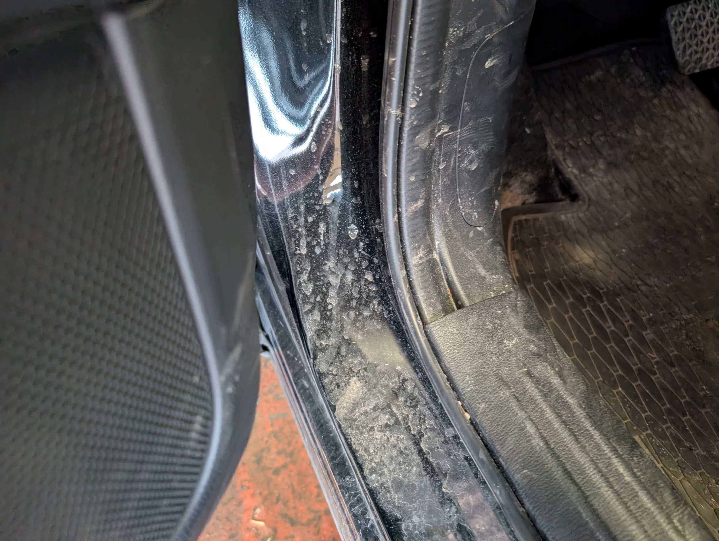 Close-up of a dirty area underneath a vehicle, showing the edge of a metal panel, the floor mat, and some dirt and grime.