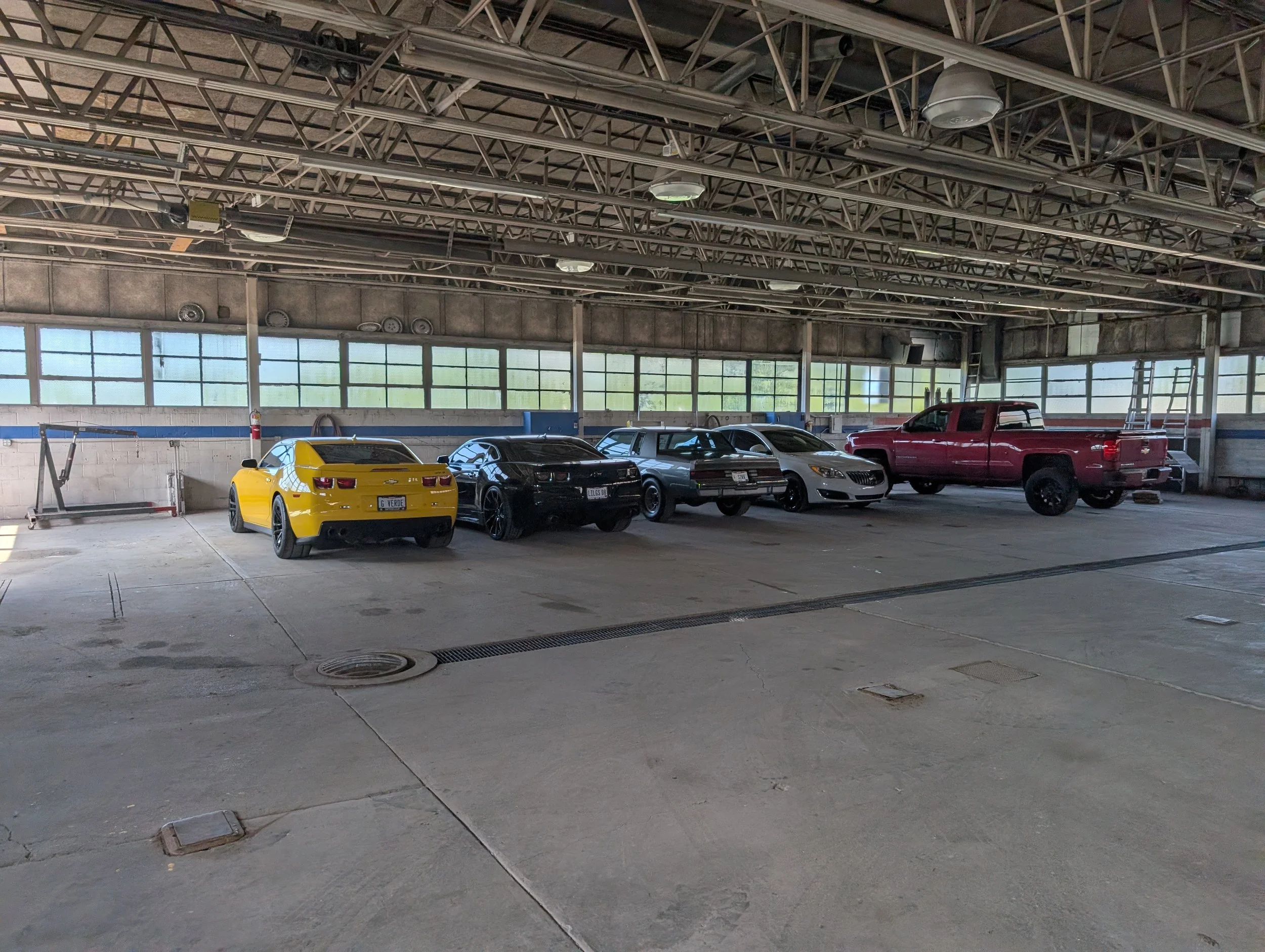 A parking garage with a yellow sports car, a black SUV, a white sedan, and a red pickup truck parked inside. The garage has a high ceiling with metal framework and large windows allowing natural light.