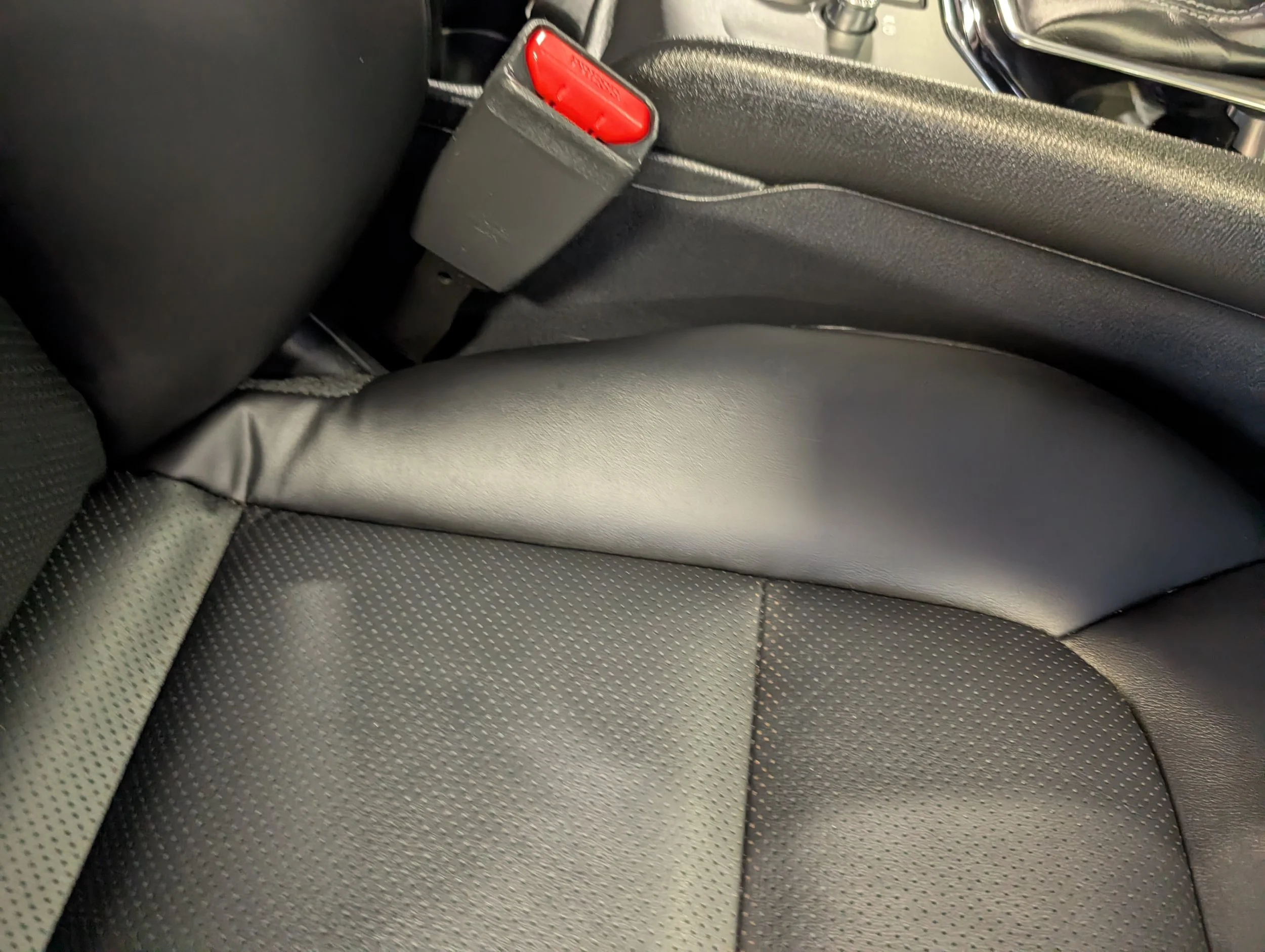 A close-up of a car's back seat showing black textured upholstery, a red seatbelt buckle, and part of the seatbelt strap.