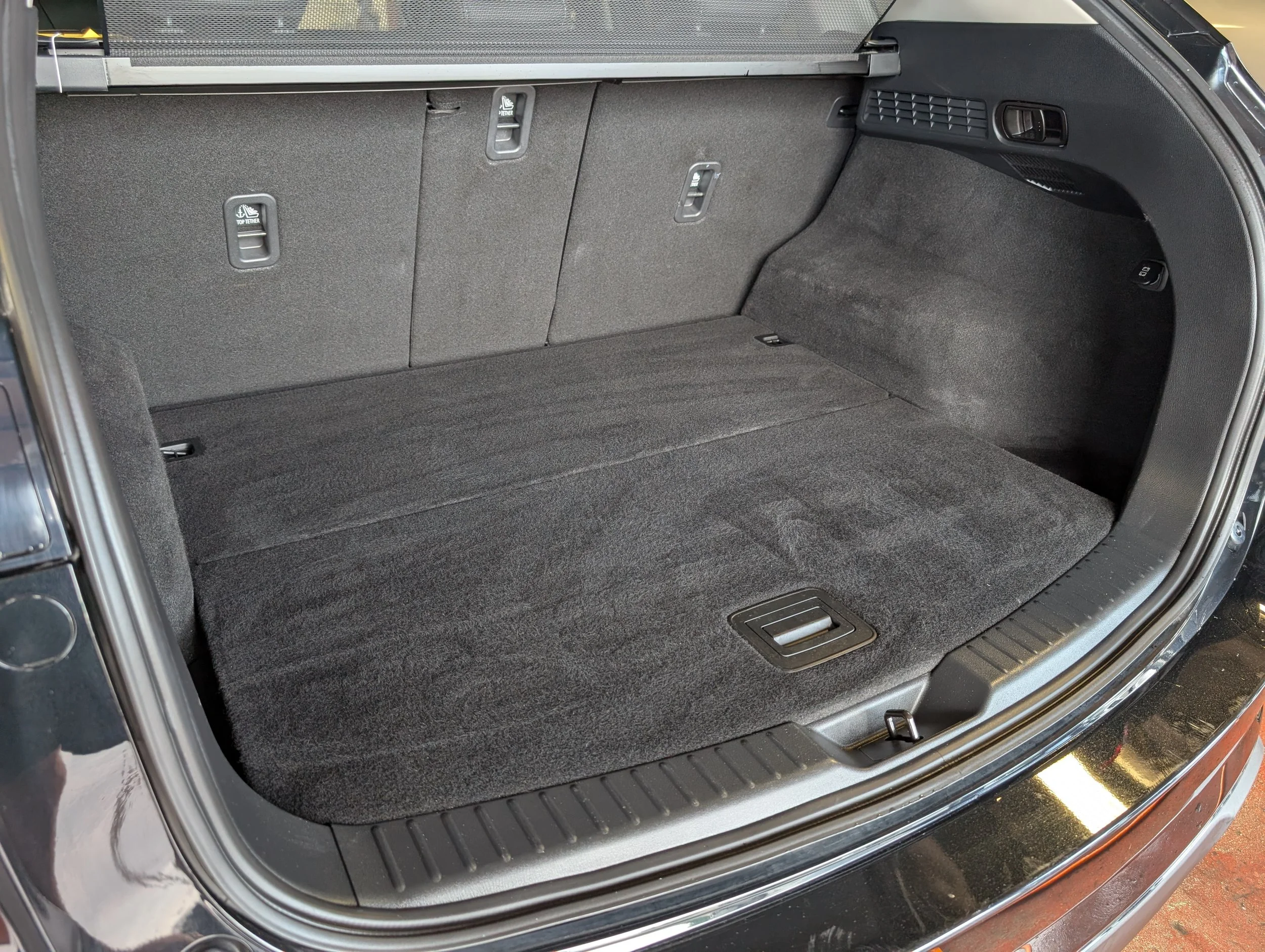 The image shows the open trunk of a vehicle with a spacious, clean, gray carpeted interior and black trim. There are three retractable tie-down hooks on the back wall of the trunk.