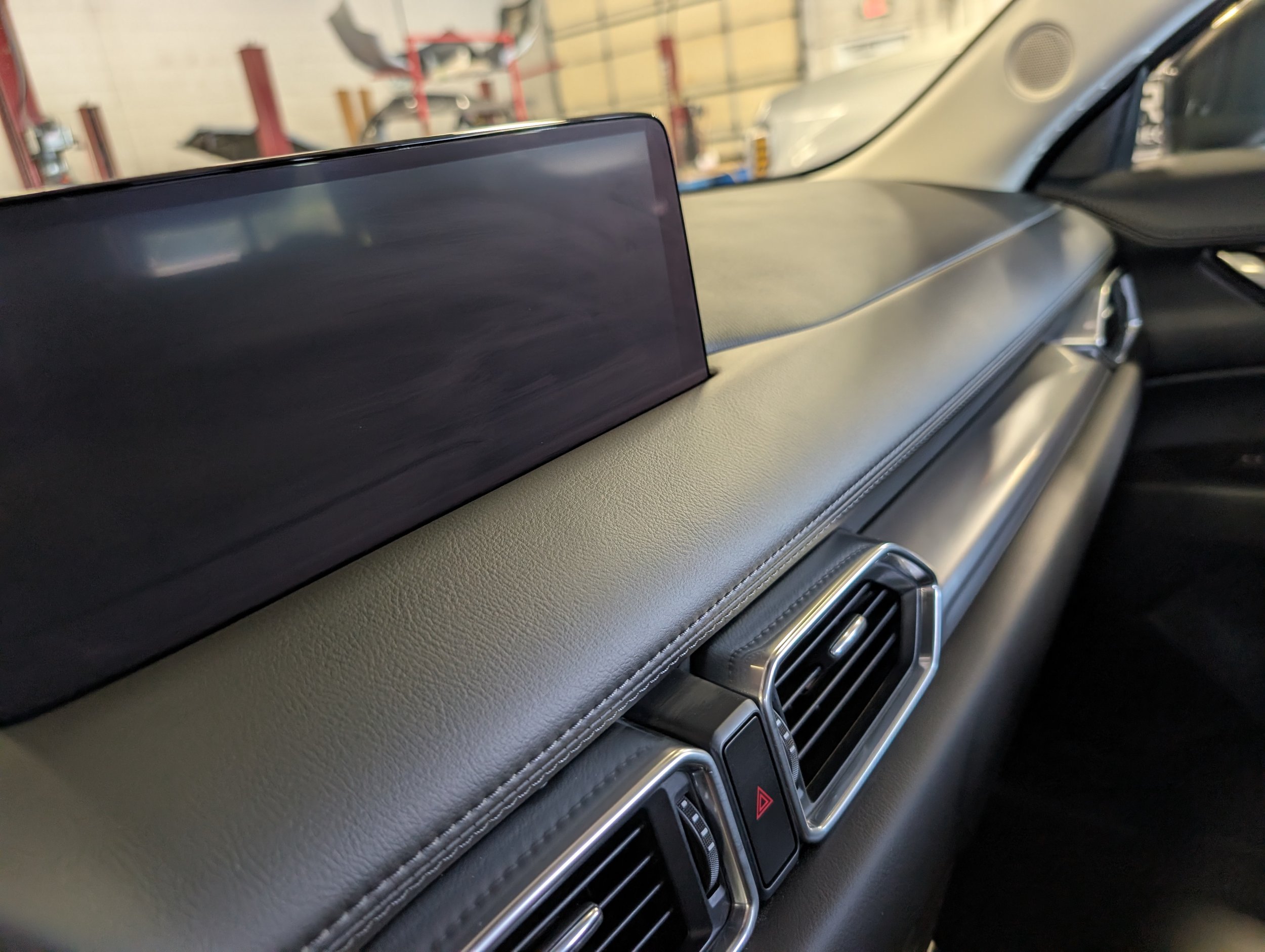 The image shows the interior dashboard of a car, focusing on the central display screen, air vents, and some controls below. The scenery outside the windshield suggests the car is parked in a garage or workshop.