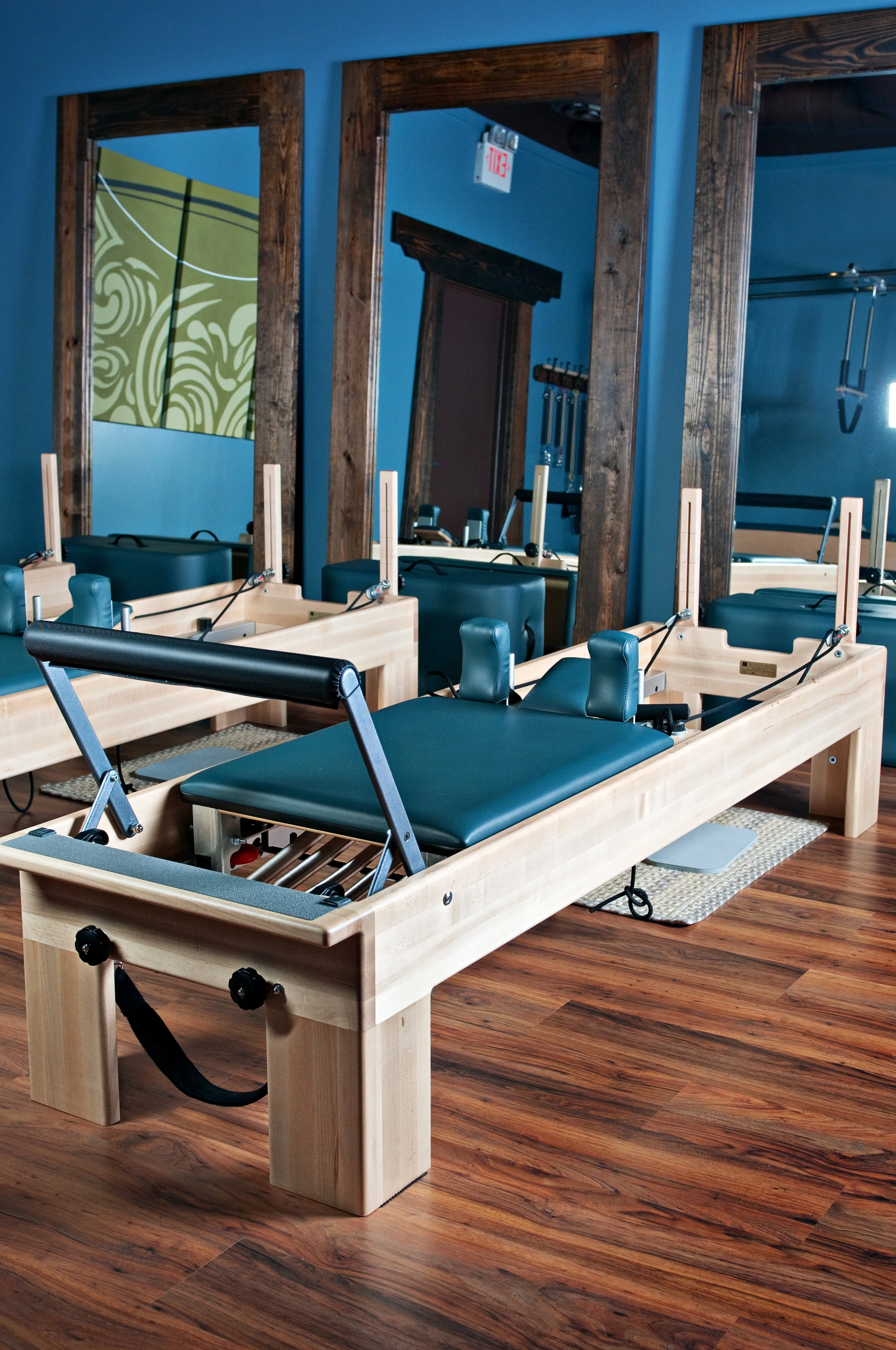 A Pilates reformer machine in a fitness studio with wooden floors, blue walls, large mirrors, and fitness equipment.