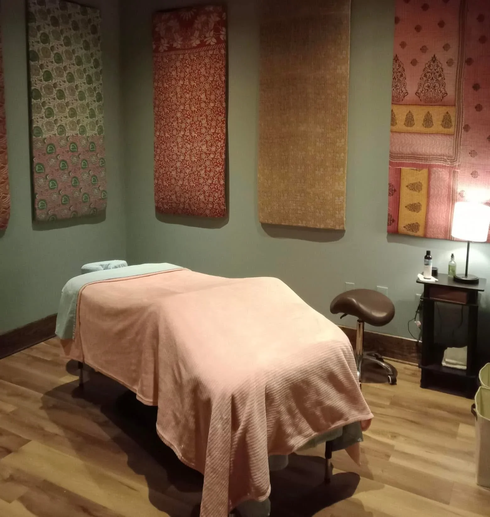 A massage therapy room with a bed covered by a pink blanket, four patterned textiles hanging on the wall, a small table with bottles and a lamp, and a stool.