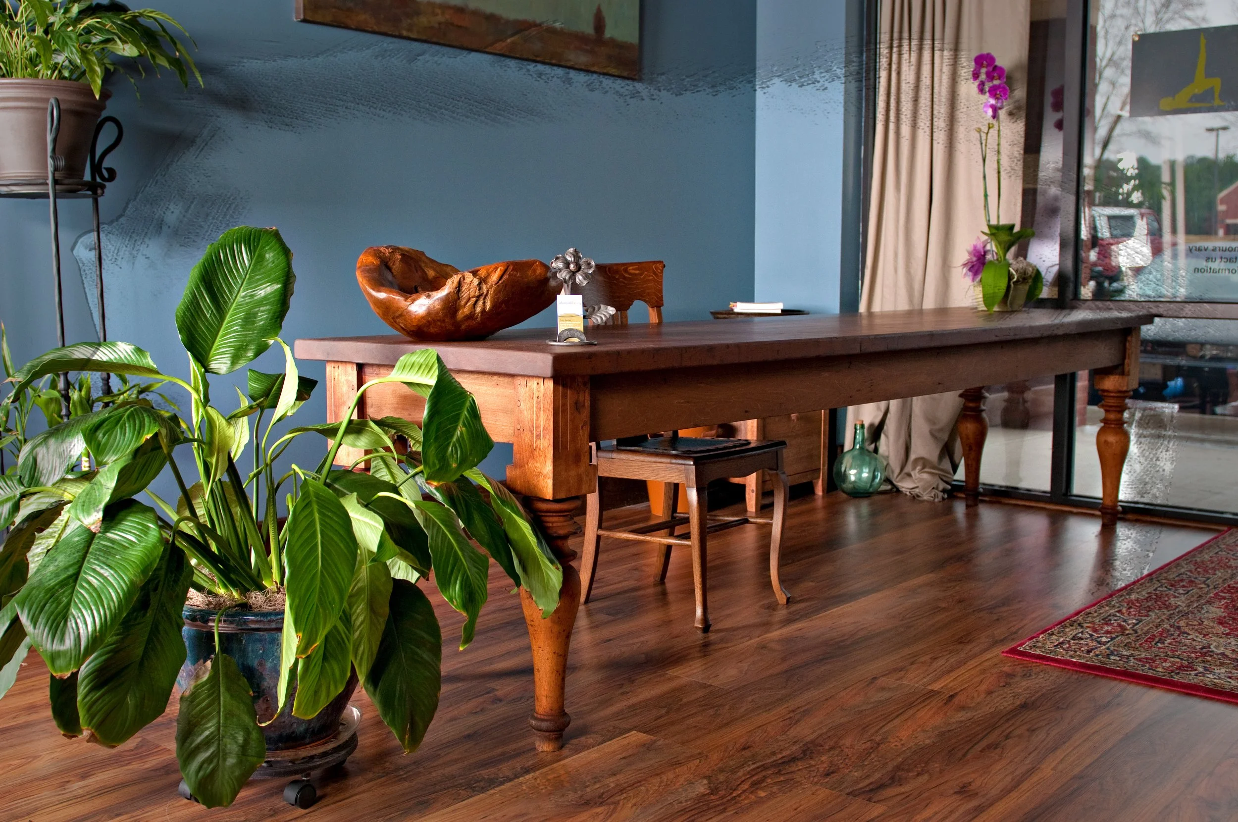 Interior of a room with a wooden table decorated with a wooden bowl and small decorative items, surrounded by potted plants, including a large leafy plant in the foreground, and featuring a glass door with curtains, a wall mirror, and colorful flowers.