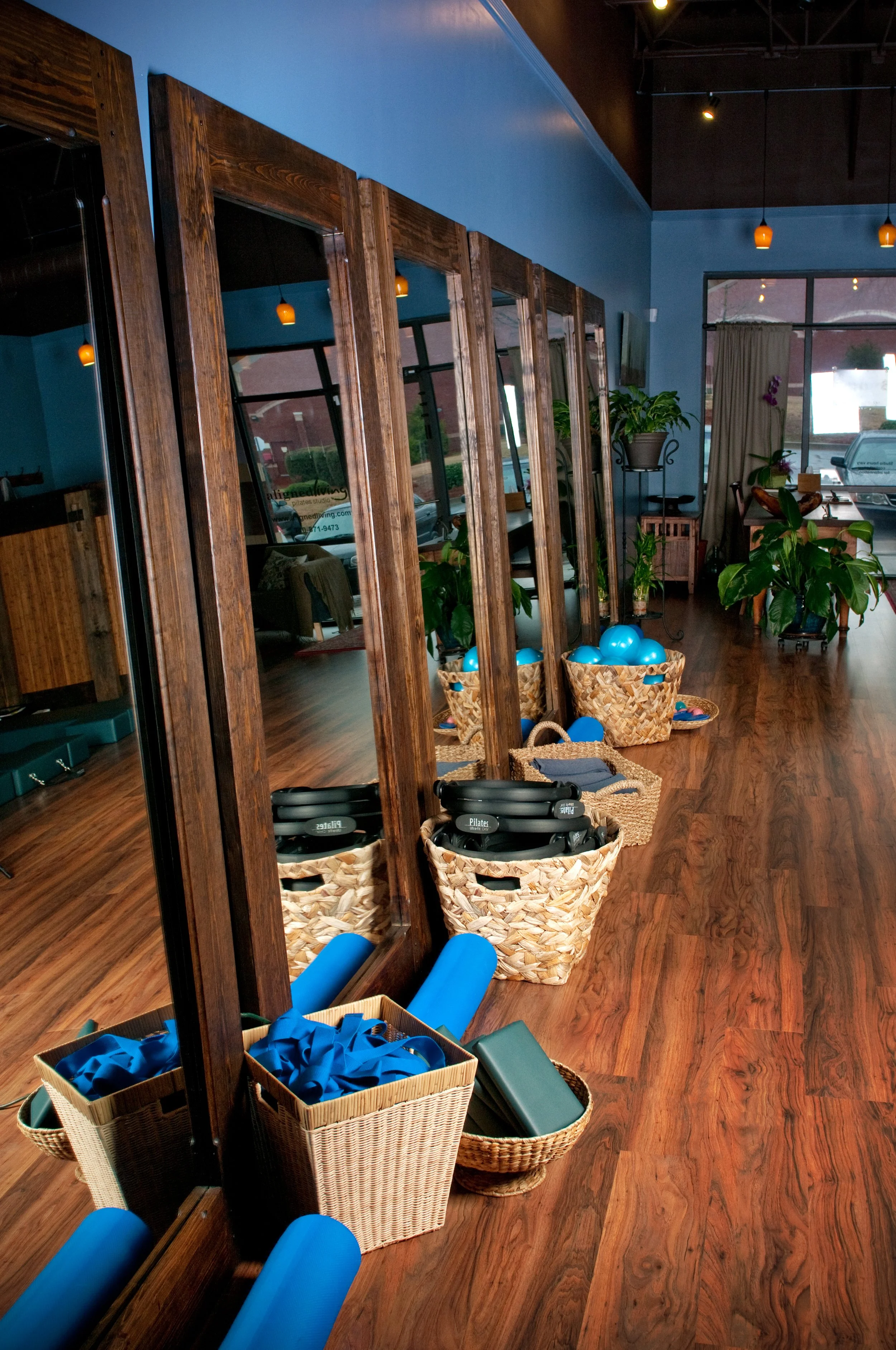 Interior of a yoga studio with mirrors, potted plants, and yoga props including blue resistance bands in baskets, foam rollers, and yoga mats on a wooden floor.