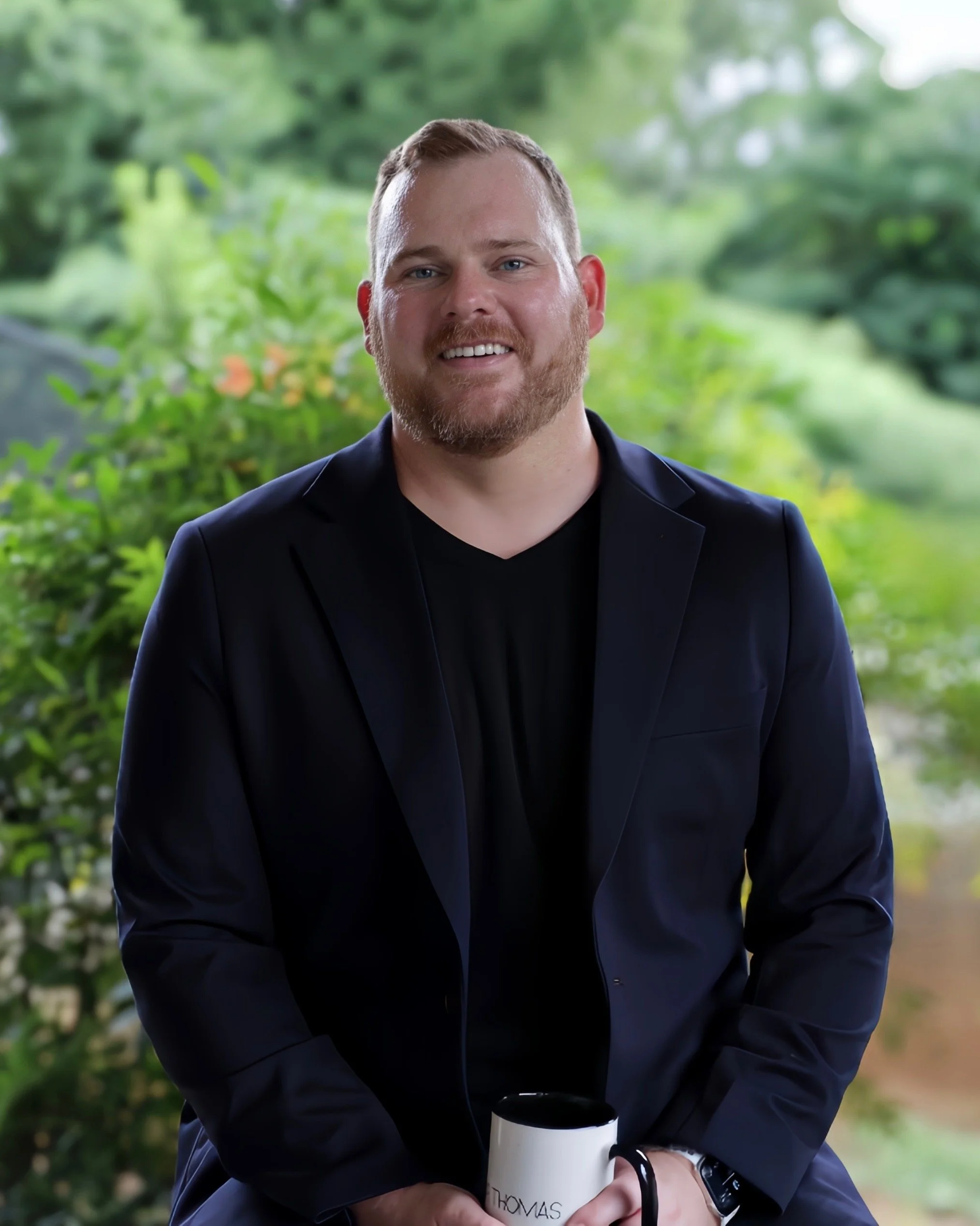 A man with a beard and short hair wearing a black blazer smiling and holding a white mug that says 'Thomas' in an outdoor setting with green foliage in the background.