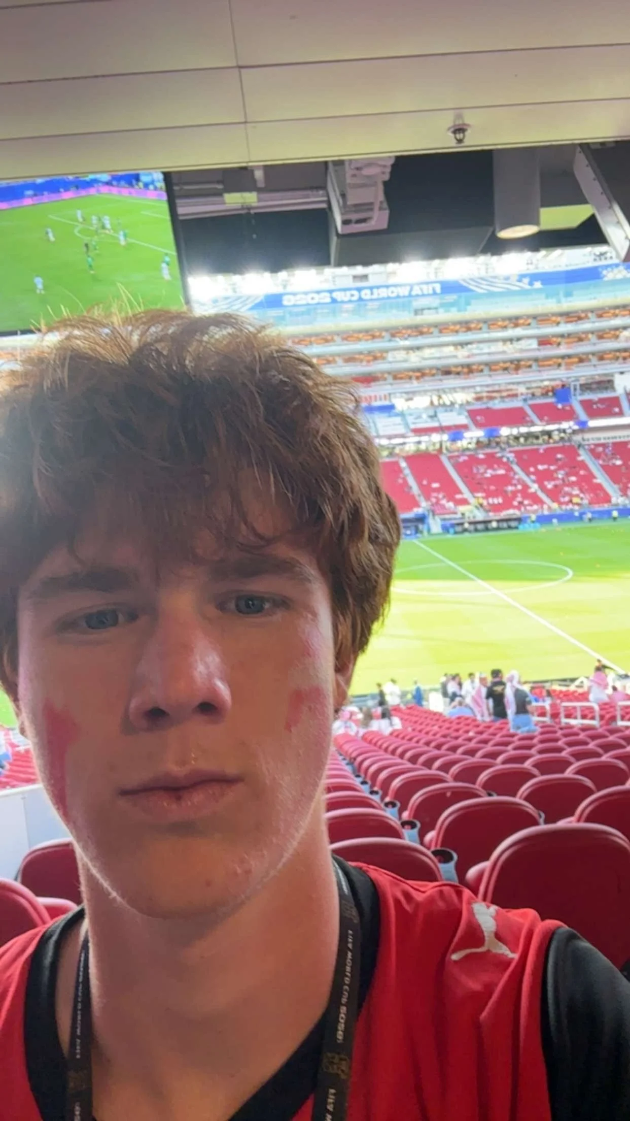 A young man with brown hair and blue eyes taking a selfie in a stadium. The background shows a soccer field with red seats, some spectators, and a large screen displaying a soccer match.