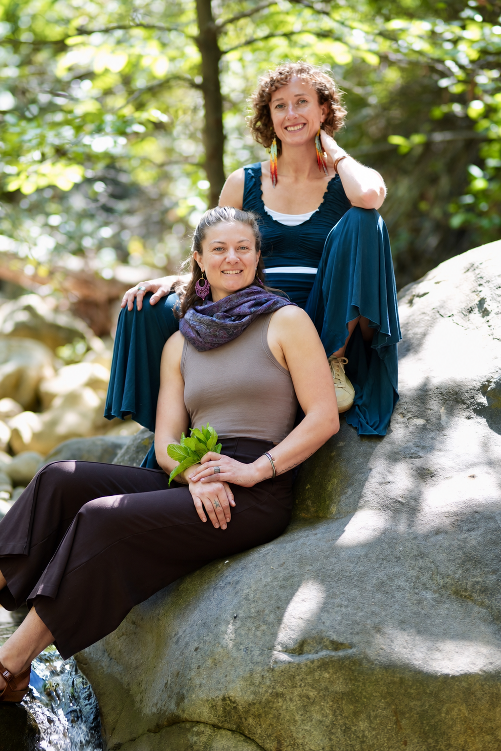 Two smiling women outdoors on a large rock in a green forest, one sitting and the other standing with one leg raised, holding a small bunch of green leaves.