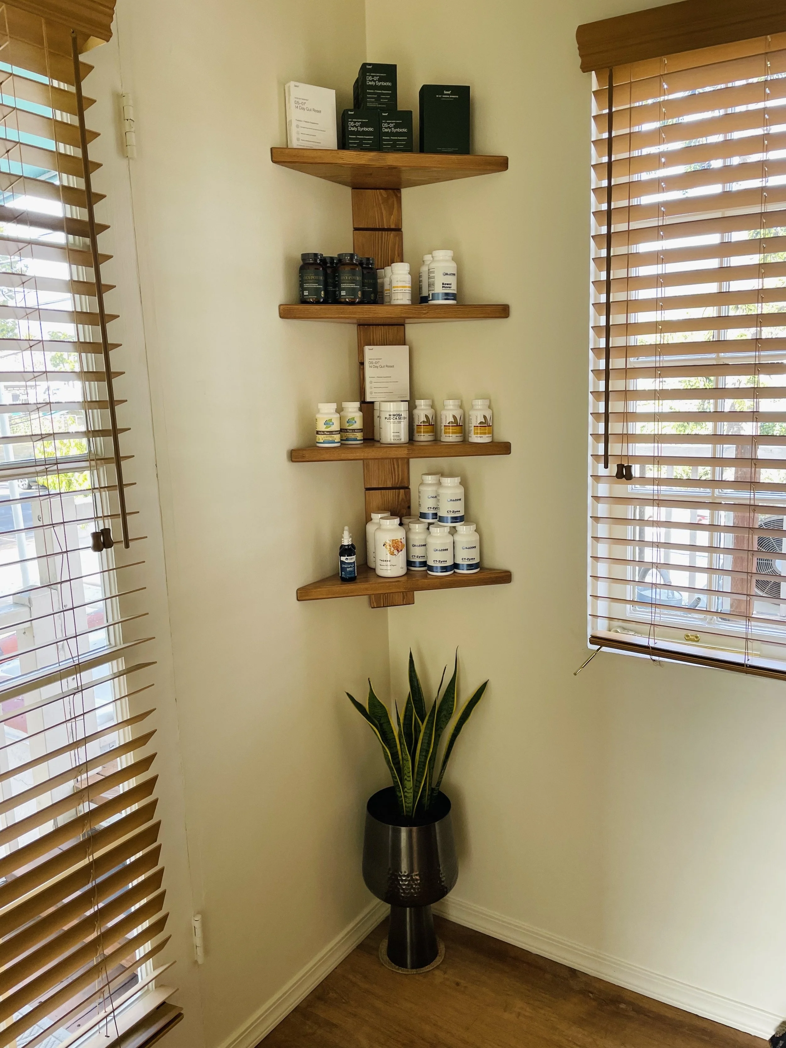 A corner of a room with beige walls, featuring a tall potted snake plant on a metal stand and a wooden corner shelf with various supplement bottles. There are wooden blinds on two windows letting in sunlight.