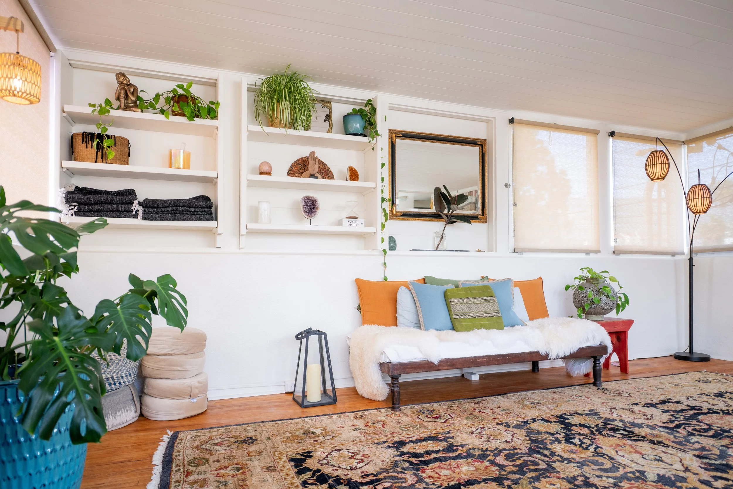 A cozy living room with a white wall, shelves decorated with plants, sculptures, and decorative objects. A mirror is hung on the wall. There are colorful pillows on a wooden bench with a fluffy throw. Two tall, artistic floor lamps are in the corner. A large patterned rug covers the hardwood floor, and leafy plants are in the foreground.
