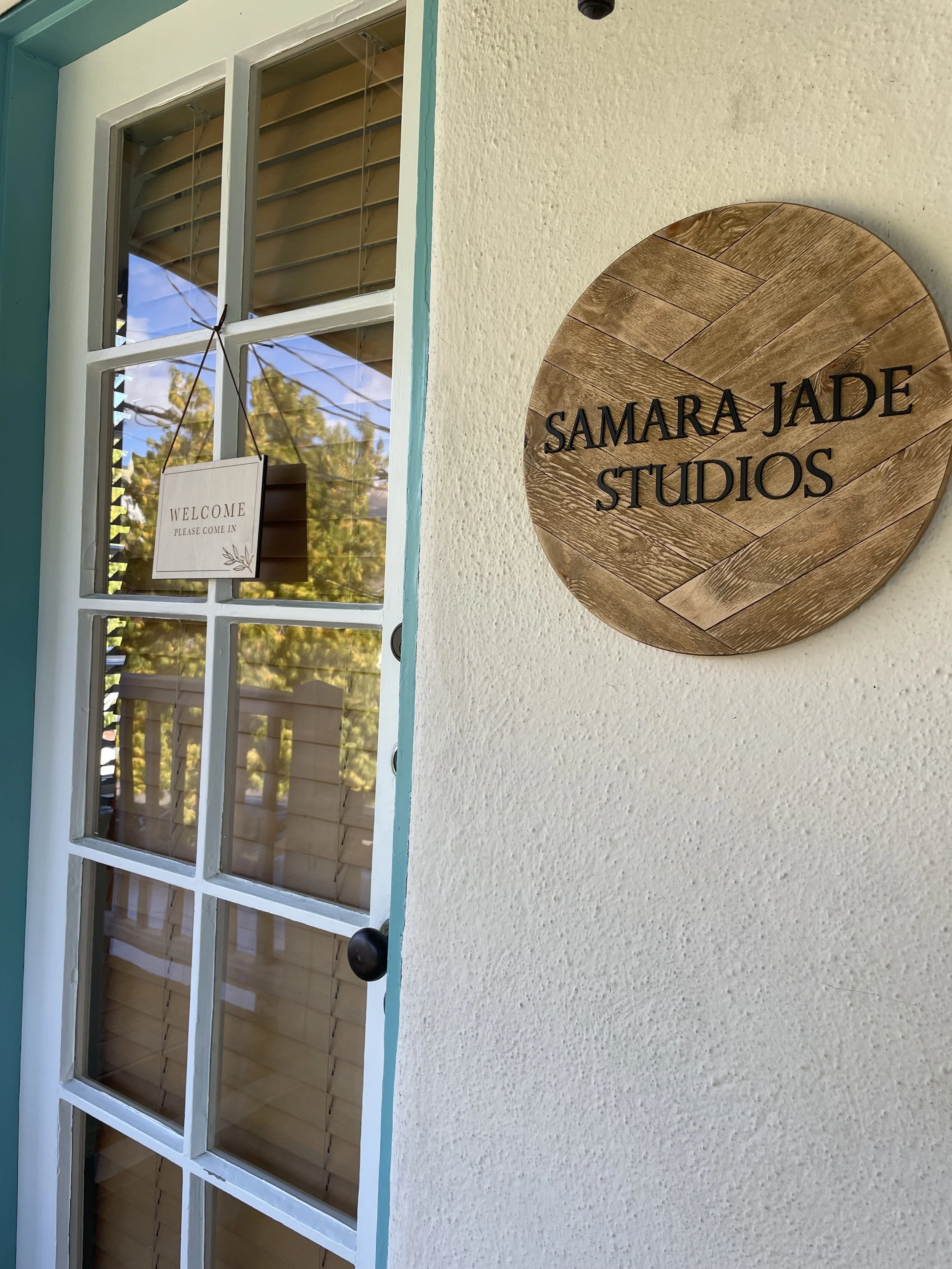 Wooden round sign on the wall reading "Samar Jade Studios" near a window with an open shutter and a sign that reads "Welcome, please come in" hanging inside