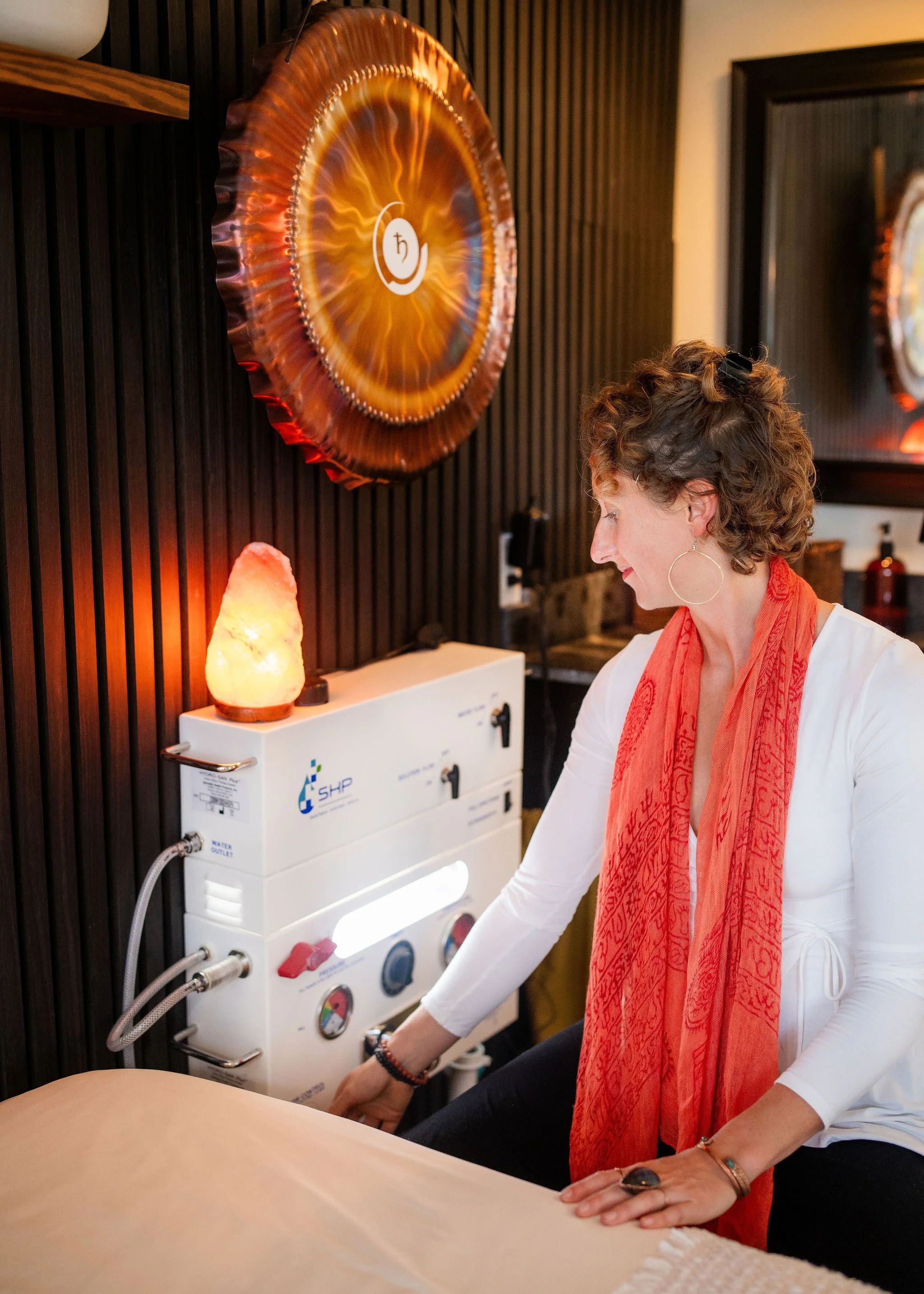 A woman with curly brown hair and hoop earrings, wearing a white shirt and red scarf, is using a machine with gauges and hoses in a room with dark wooden walls. There is a salt lamp on a table near her and framed artwork on the wall.