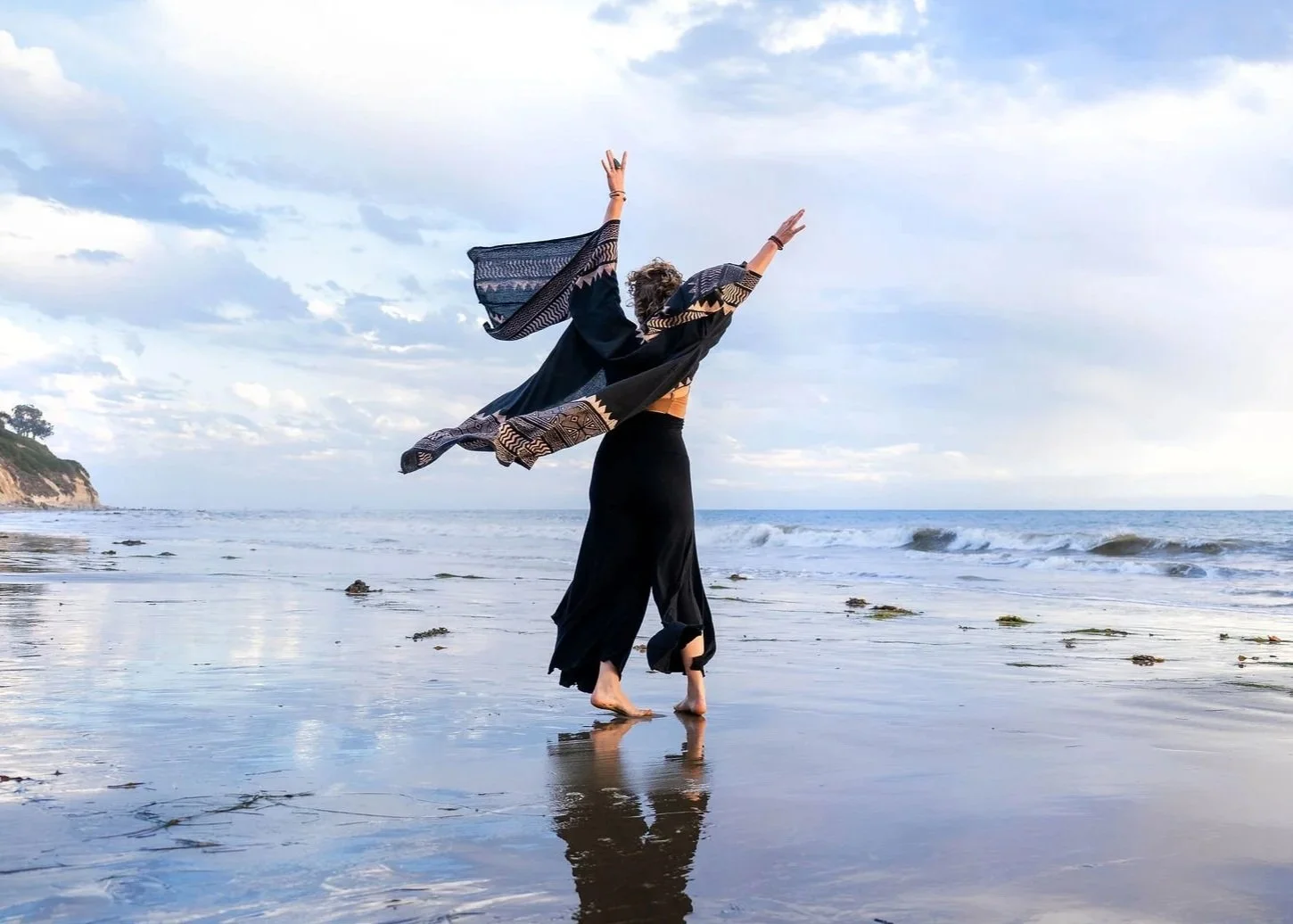 A person dancing or performing on the beach near the ocean, with arms raised, wearing a black skirt and a patterned shawl or scarf, with a cloudy sky in the background.