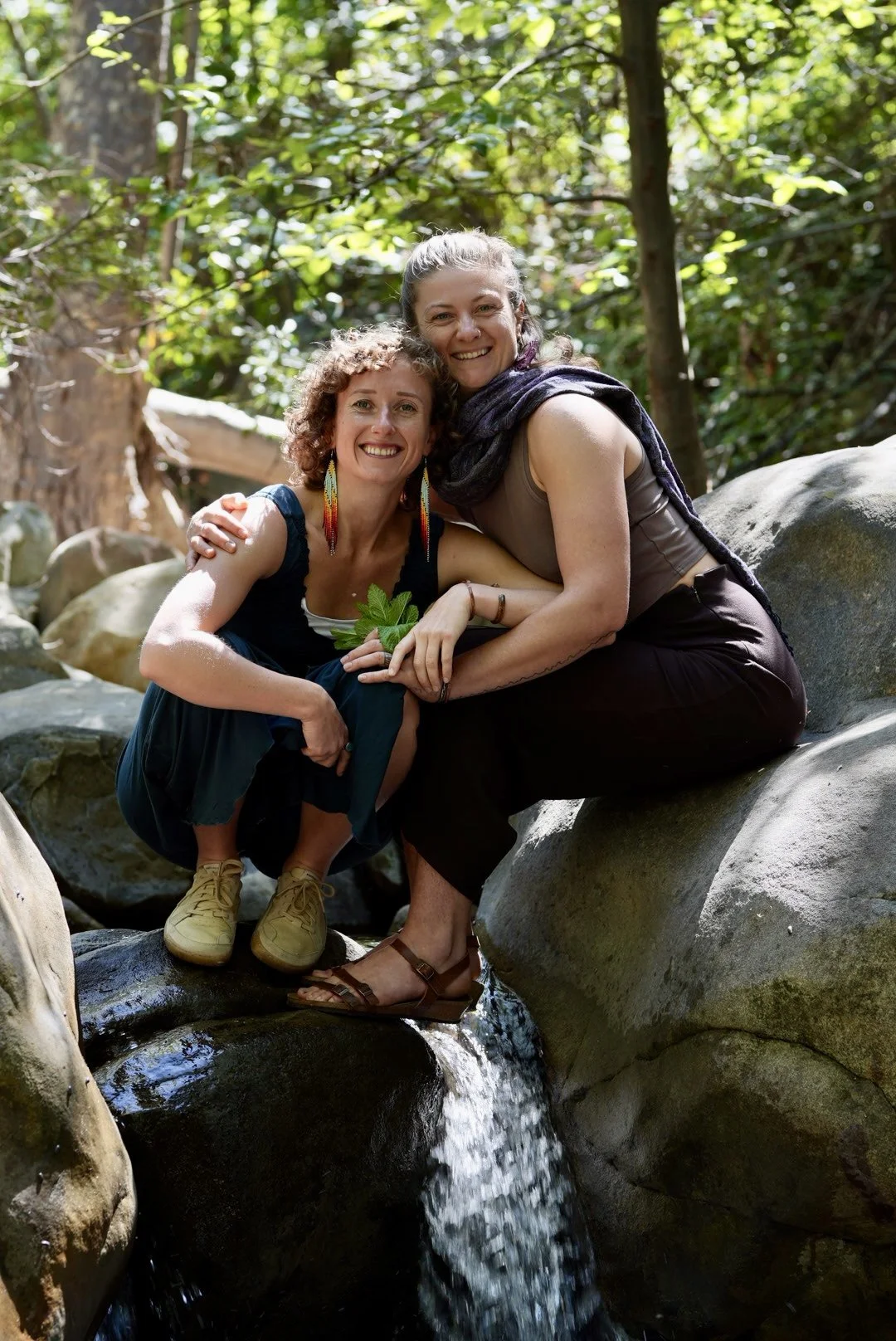 Two women sitting on rocks beside a small stream in a forest, smiling and embracing each other, surrounded by green foliage.