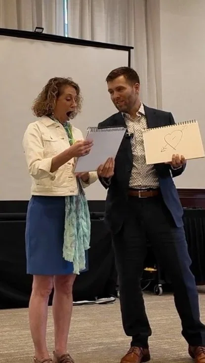 A woman and a man are standing together, holding sketchbooks, with the woman showing surprise and the man holding a drawing of a heart with an arrow.
