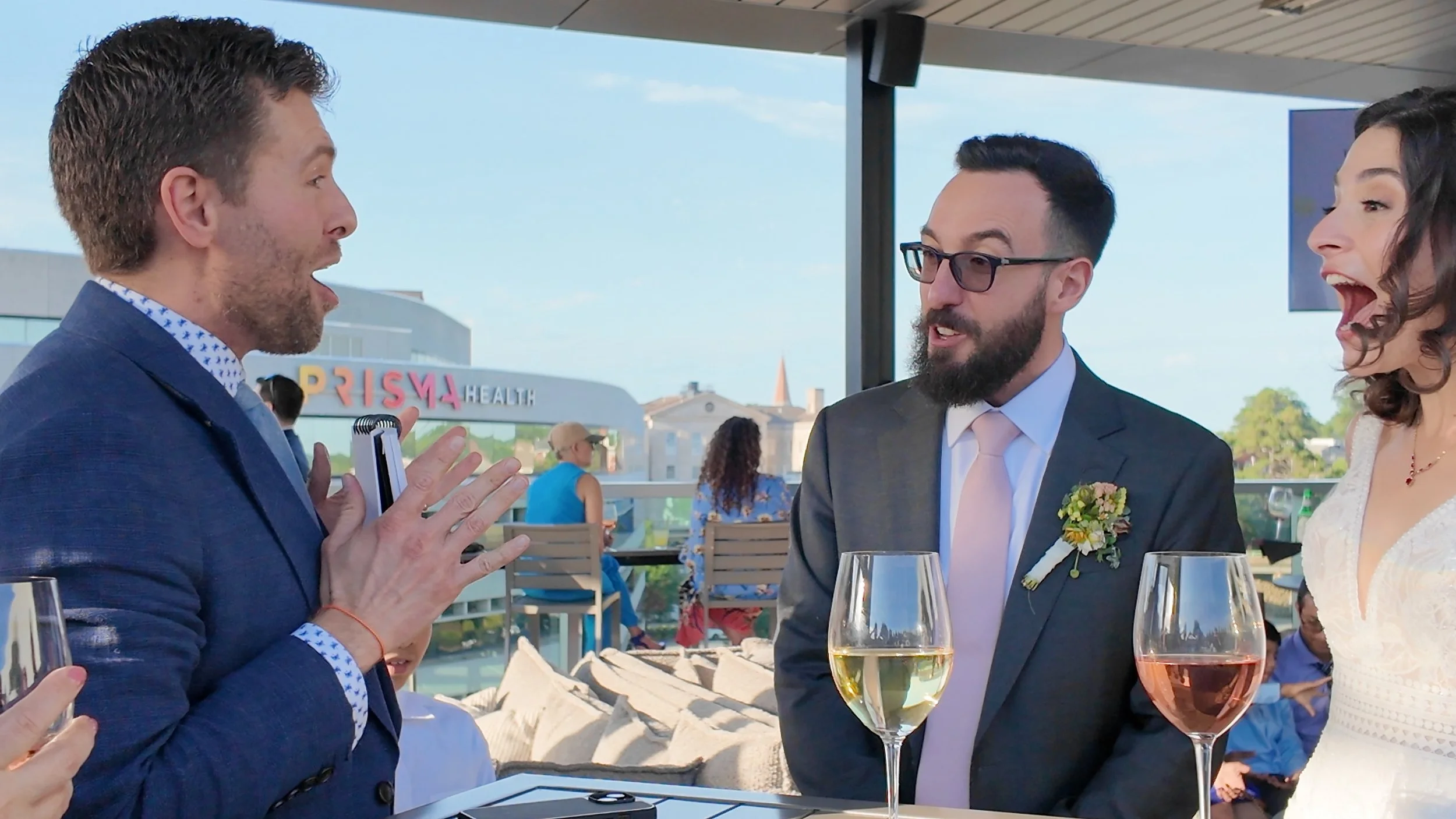 Three people, two men and a woman, engaged in conversation during a wedding reception on a rooftop patio. They are delighted by a magic trick.