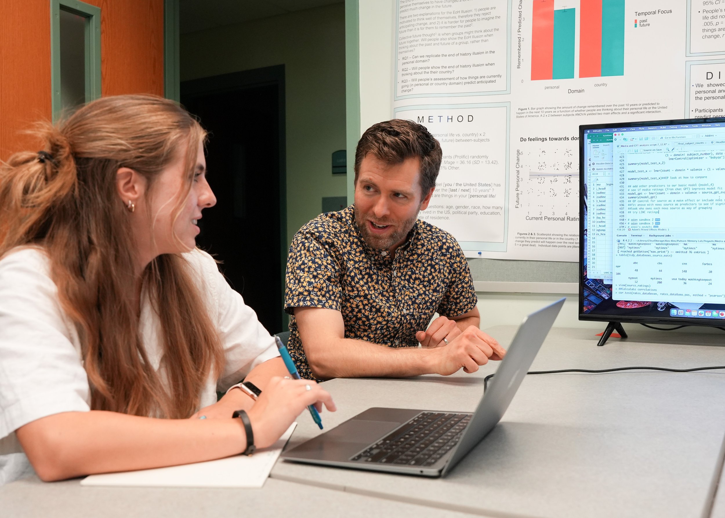 Two people, a woman with long red hair and a man with dark hair, are sitting at a table with laptops, engaged in a discussion in a conference room. They are surrounded by printed research posters and a computer screen displaying data analysis.