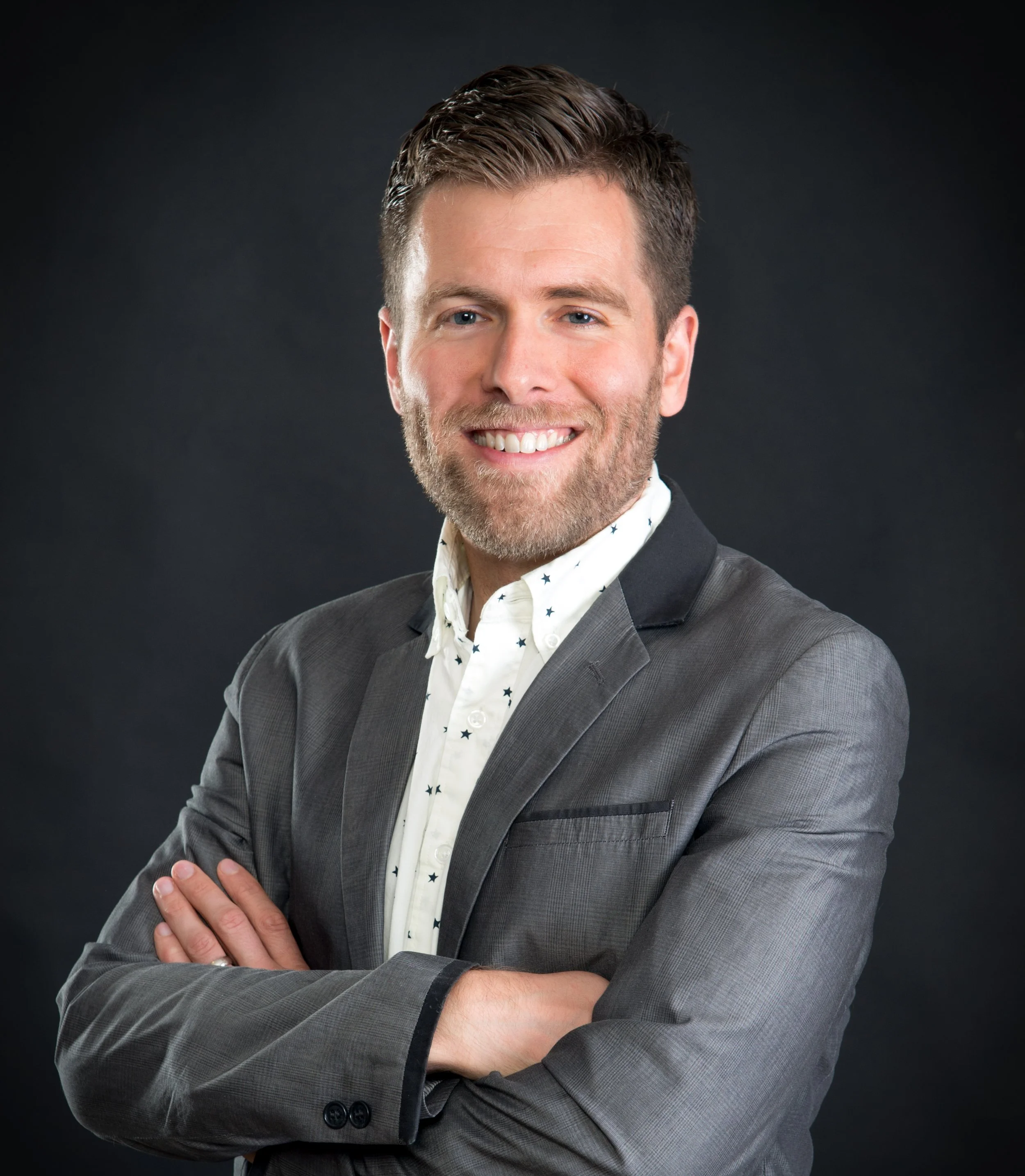 A smiling man wearing a dark gray suit jacket and a white shirt with small star patterns, posing against a dark background.