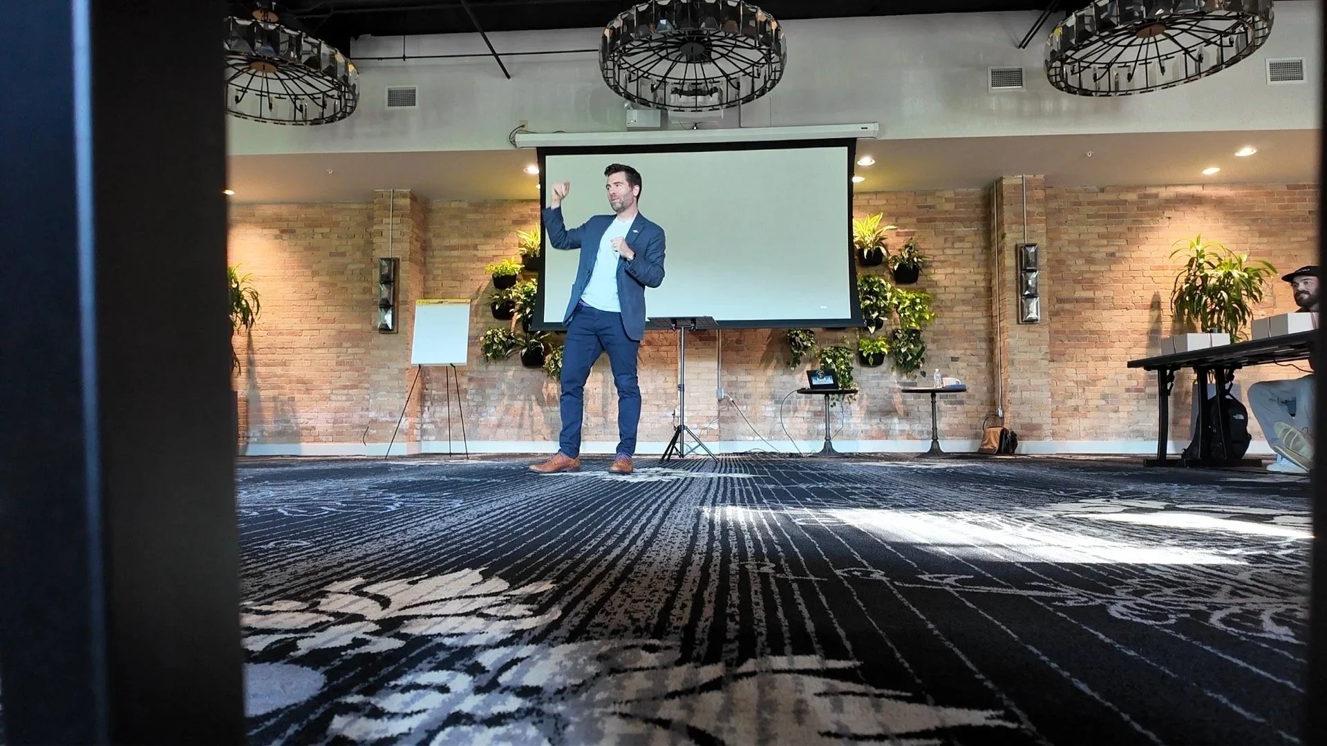 A man in a blue suit giving a presentation in a conference room with a large screen behind him, potted plants on the wall, and a patterned carpet on the floor.