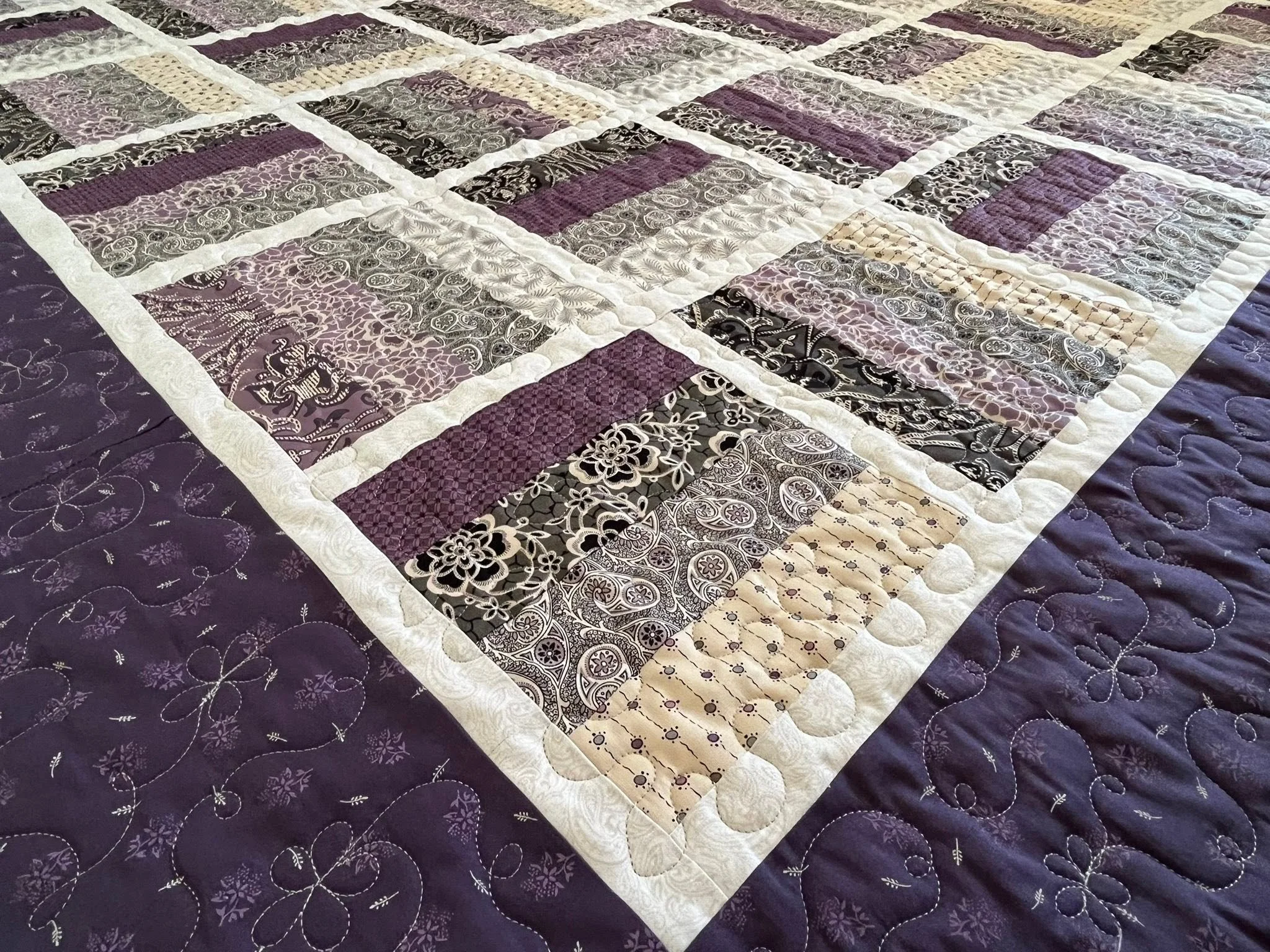 A quilt with a pattern of purple, black, beige, and lavender squares featuring floral and abstract designs, sewn with white borders.